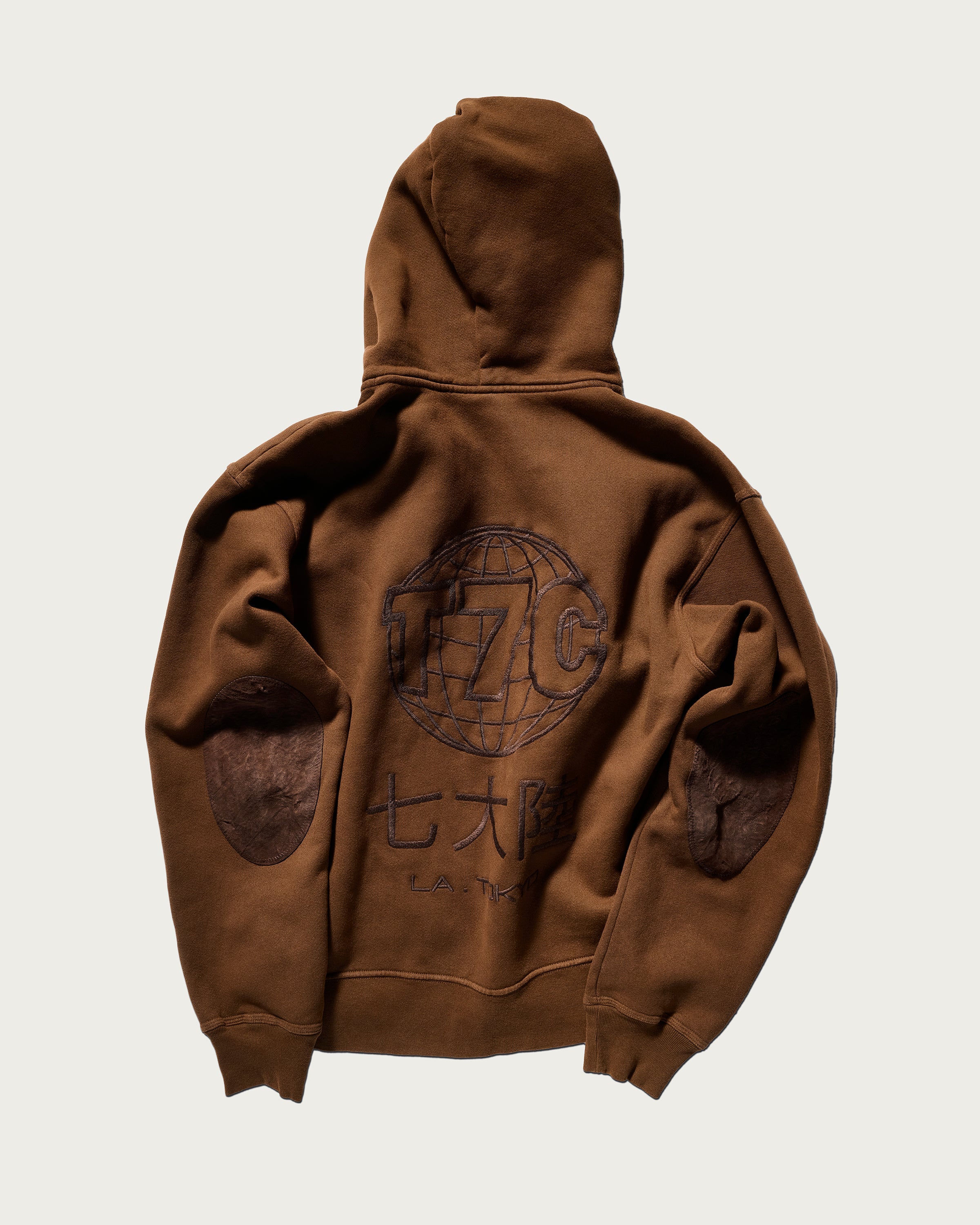 The Camden Hoodie in Pecan by T7C The Seven Continents features a relaxed fit, suede elbow patches, and an embroidered globe with “T7C,” Japanese characters, and “L.A. TOKYO” on the back. Shown laid flat on a light background.