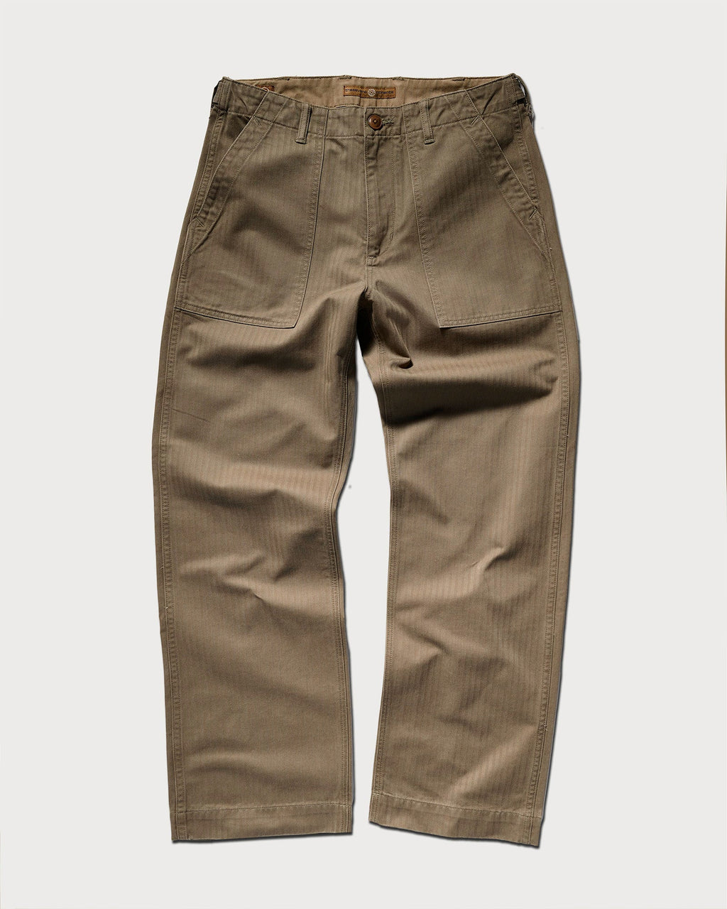 The T7C Liam Military Pant in Olive Drab features a relaxed fit, large front patch pockets, belt loops, and a button closure, all crafted from cotton herringbone fabric and shown laid flat on a white background.