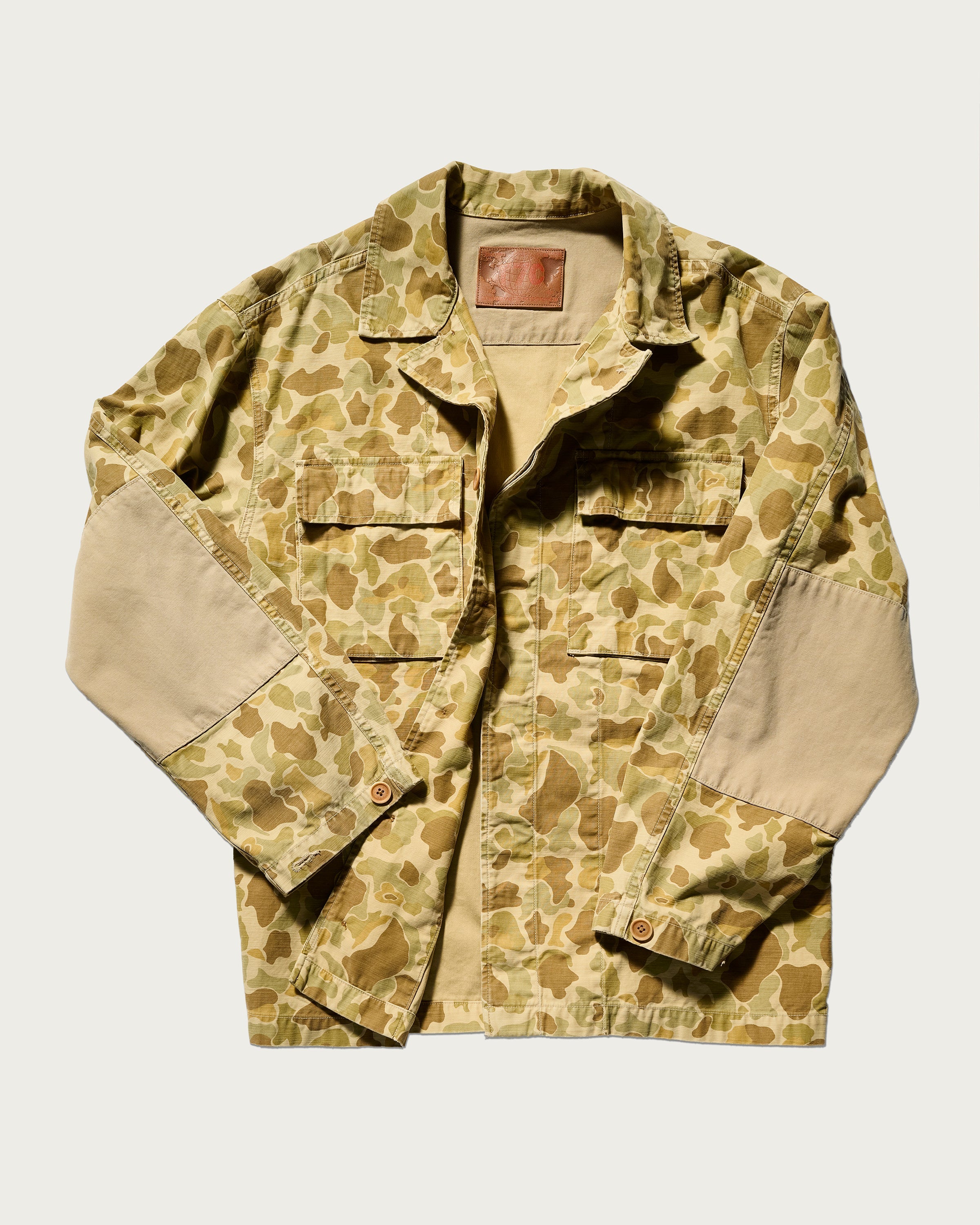 The Ryan Jacket in Camouflage by T7C The Seven Continents features a light brown camo print, two chest pockets, tan elbow patches, and a brown leather inner collar patch in relaxed-fit cotton-back satin, shown on a white background.