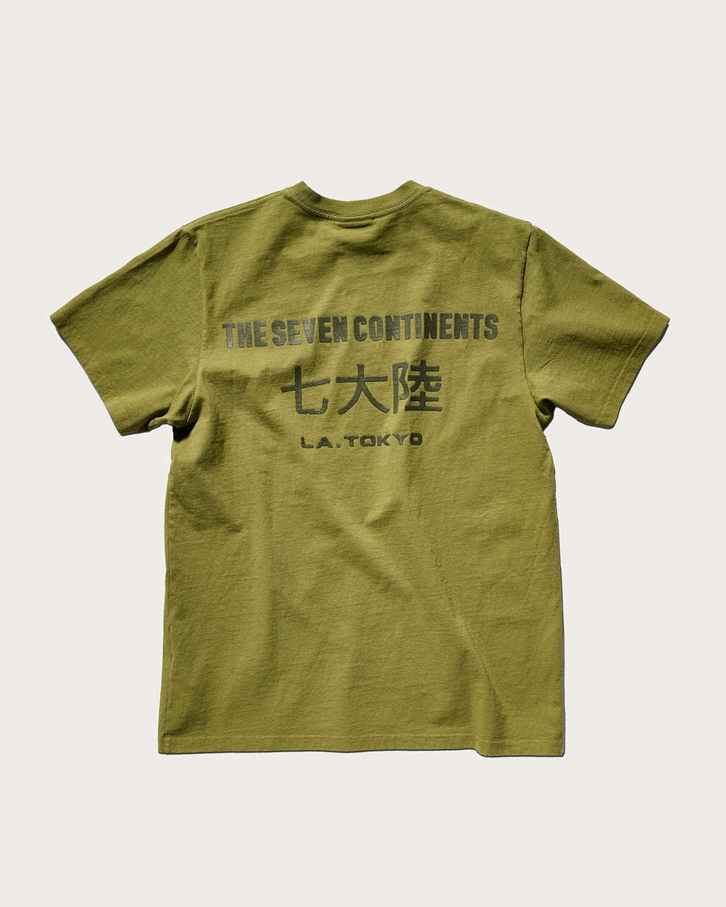 The Timo Tee in Olive by T7C The Seven Continents features extra softness and a dark gray puff print on the back with “THE SEVEN CONTINENTS,” Japanese characters, and “L.A. TOKYO.” Laid flat on a white background.