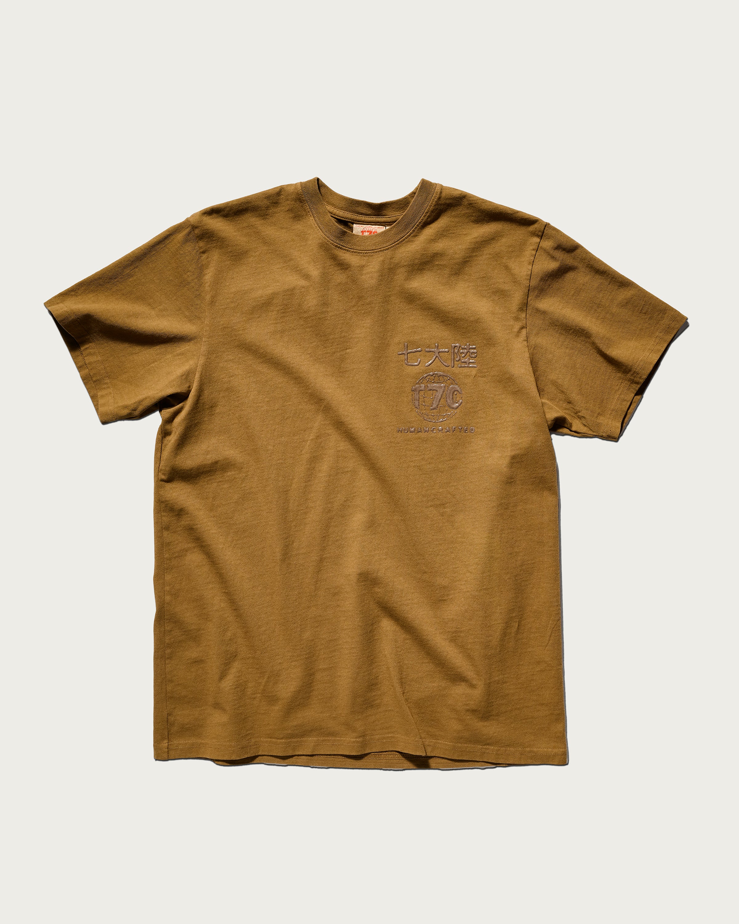 The Timo Tee in Tobacco by T7C The Seven Continents is a soft, olive green short-sleeve t-shirt featuring subtle embroidered text and symbols on the left chest, displayed flat on a white background.