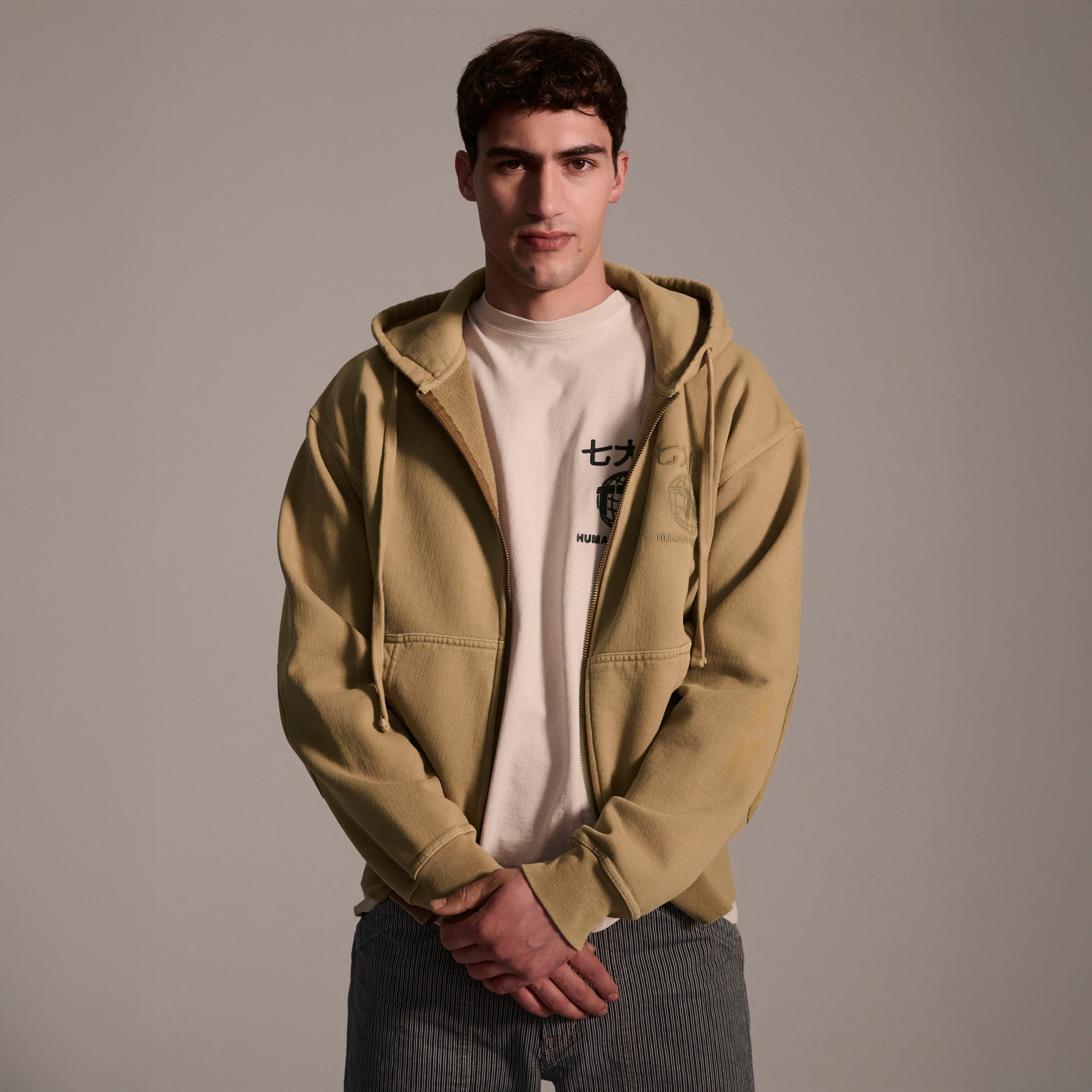 A young man with short dark hair stands against a light gray background, wearing T7C The Seven Continents Camden Hoodie in Desert over a white T-shirt and gray striped pants, with his hands clasped in front of him.