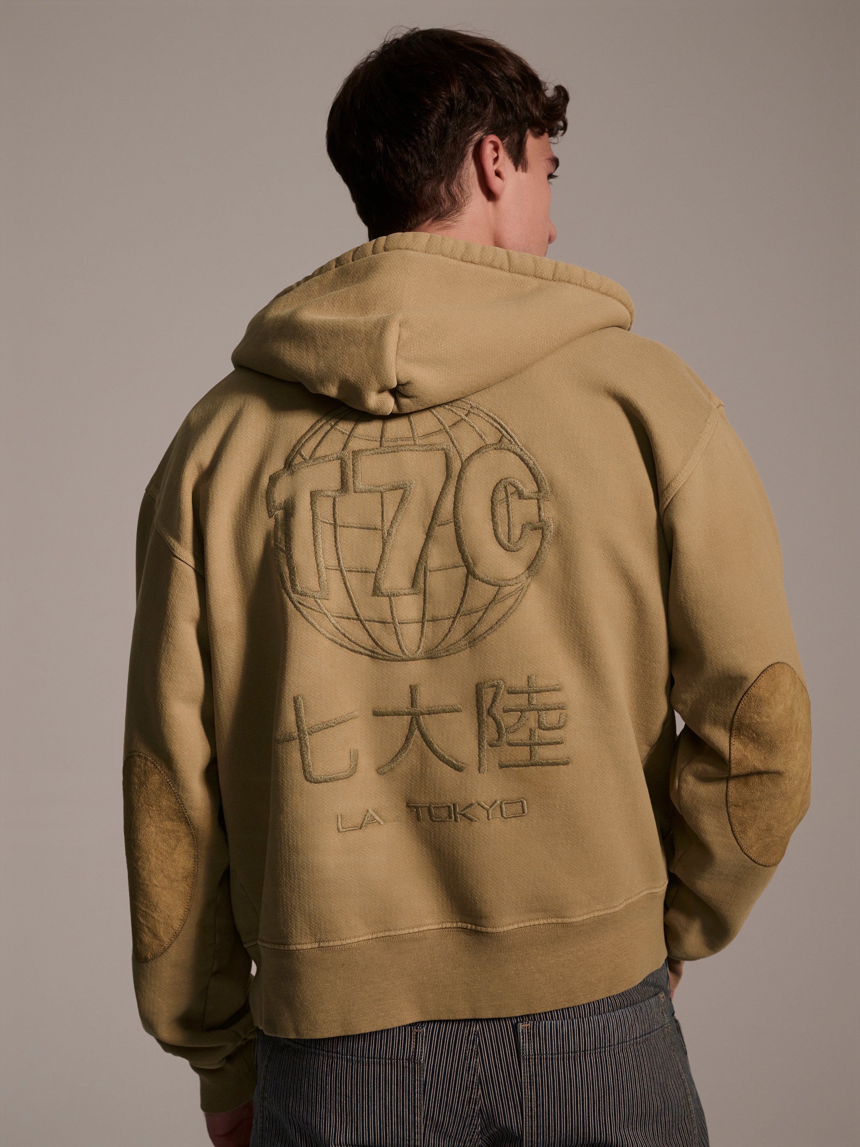A person faces away from the camera in The Camden Hoodie in Desert by T7C The Seven Continents, featuring a large globe graphic, Japanese characters, LA TOKYO on the back, and suede elbow patches for a stylish, relaxed vibe.