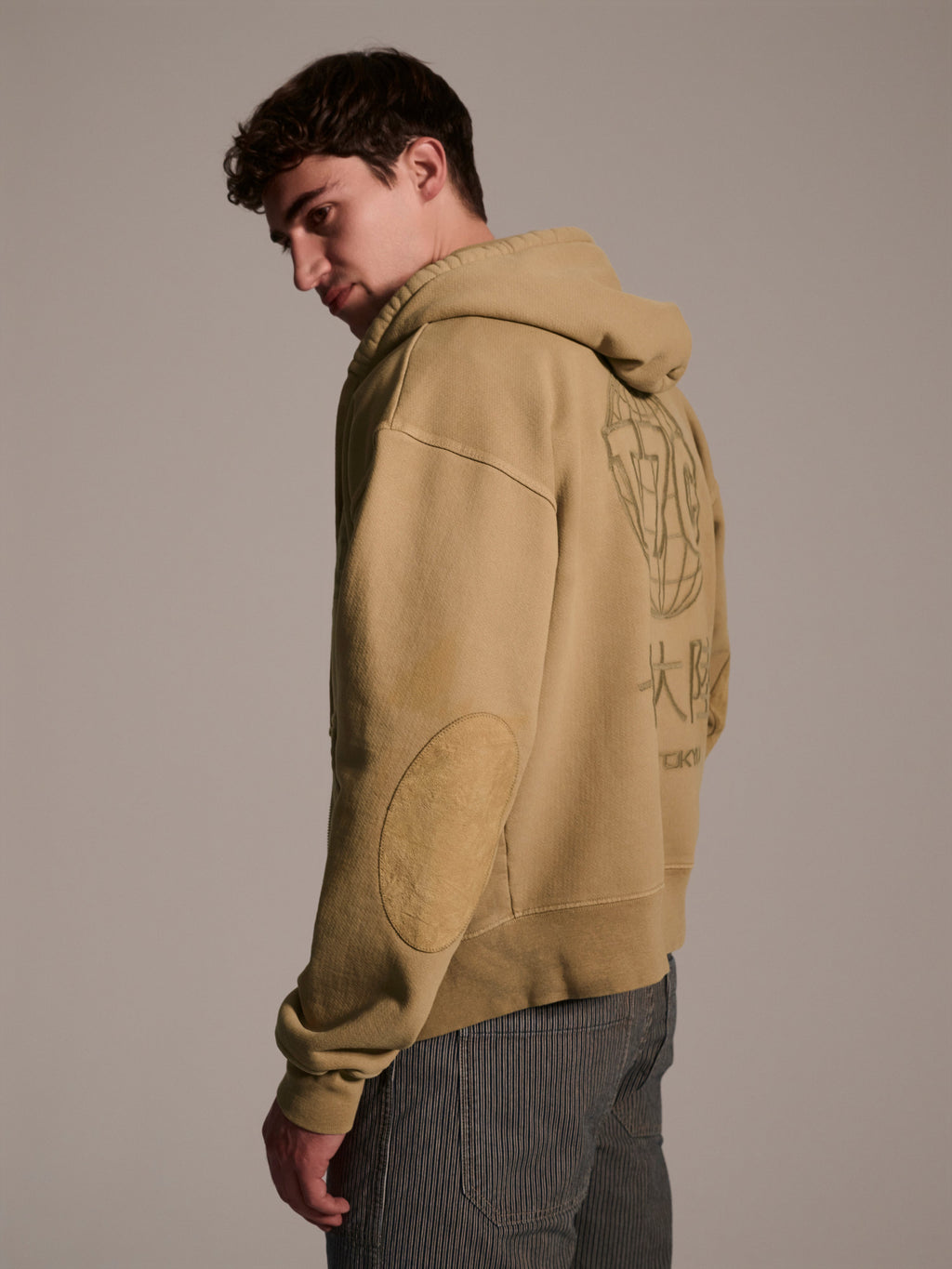A young man with short brown hair wears The Camden Hoodie in Desert by T7C The Seven Continents, featuring back graphics and suede elbow patches. He pairs it with striped pants and stands looking over his shoulder against a plain background.