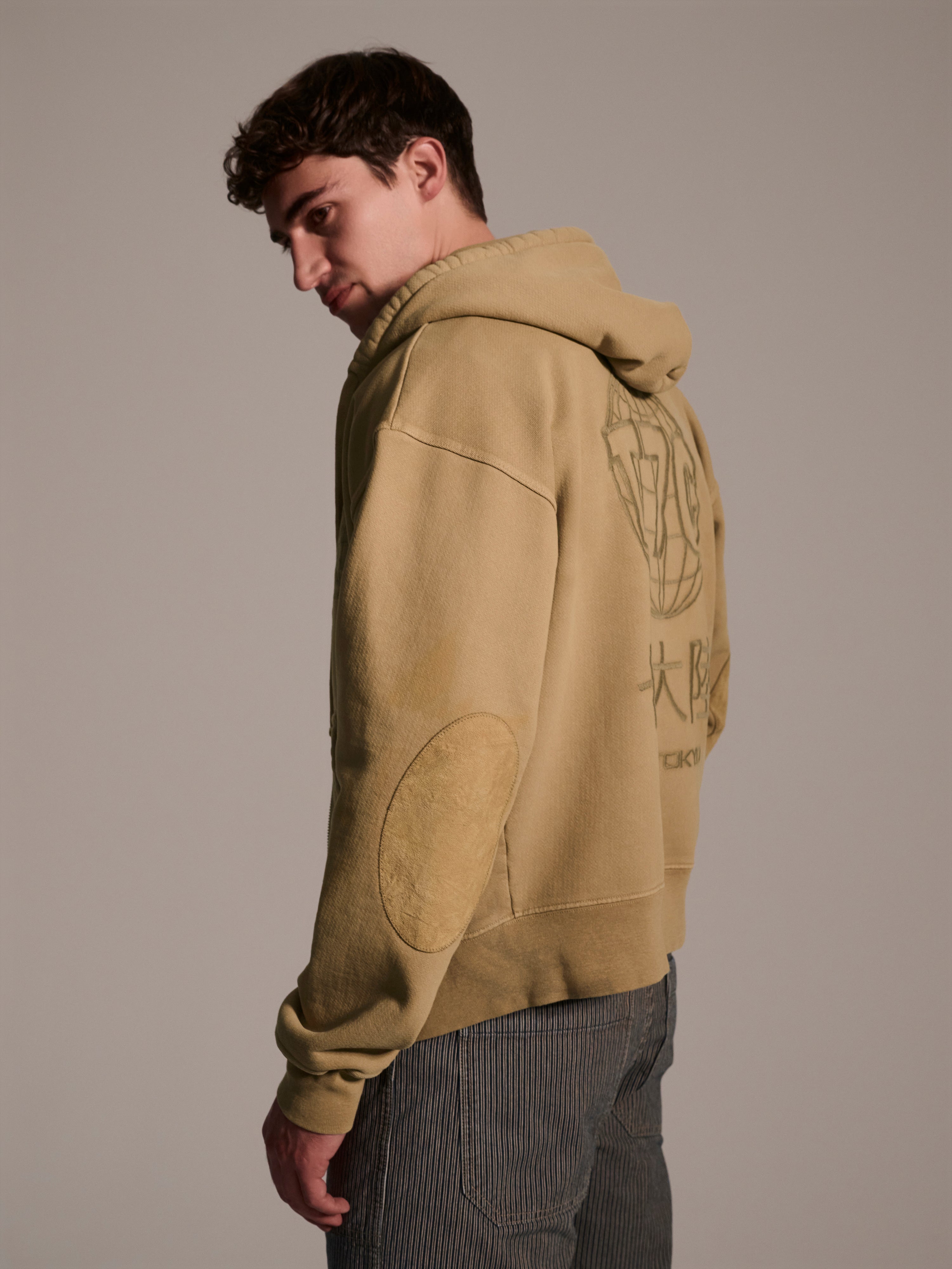 A young man with short brown hair wears The Camden Hoodie in Desert by T7C The Seven Continents, featuring back graphics and suede elbow patches. He pairs it with striped pants and stands looking over his shoulder against a plain background.