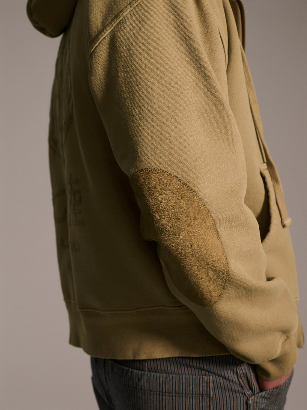 A person wears The Camden Hoodie in Desert by T7C The Seven Continents, featuring suede elbow patches, and blue pinstriped pants, standing sideways with one hand in their pocket against a neutral background.