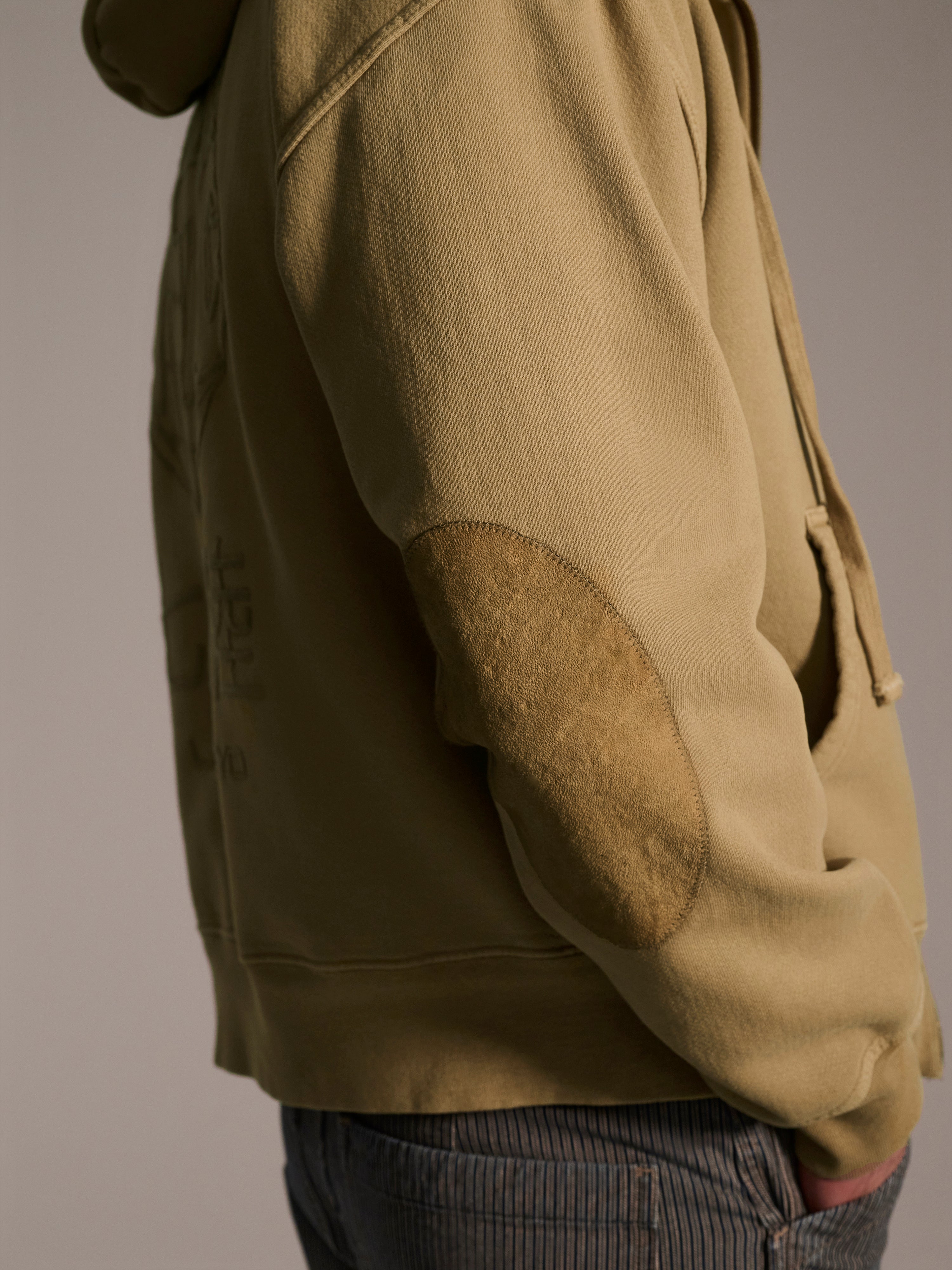 A person wears The Camden Hoodie in Desert by T7C The Seven Continents, featuring suede elbow patches, and blue pinstriped pants, standing sideways with one hand in their pocket against a neutral background.
