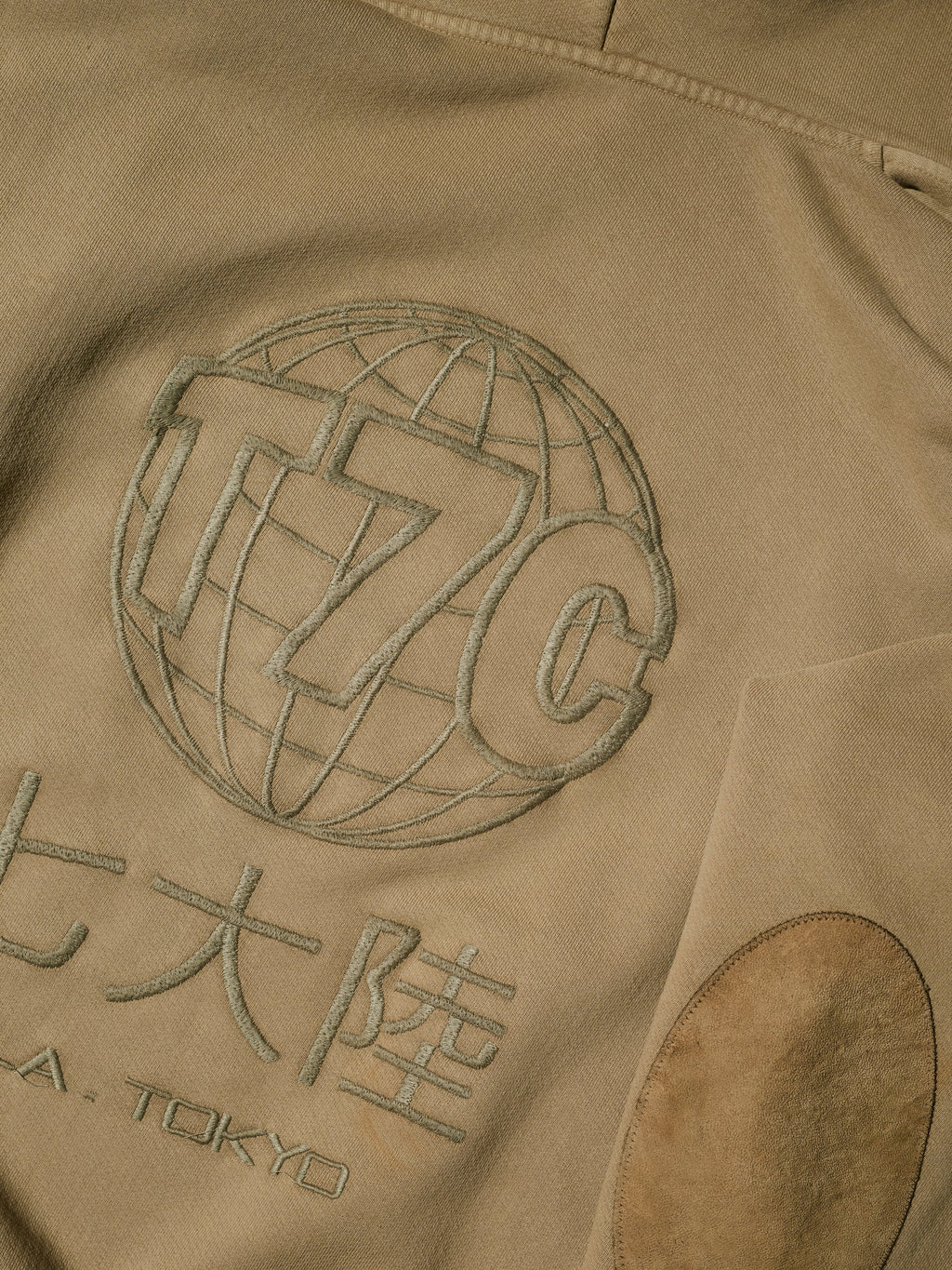 A close-up of tan fabric with embroidered details—a globe with “T7C,” Japanese characters, and “A. TOKYO” underneath. The Camden Hoodie in Desert by T7C The Seven Continents includes an oval sleeve patch and suede elbow patches for style.