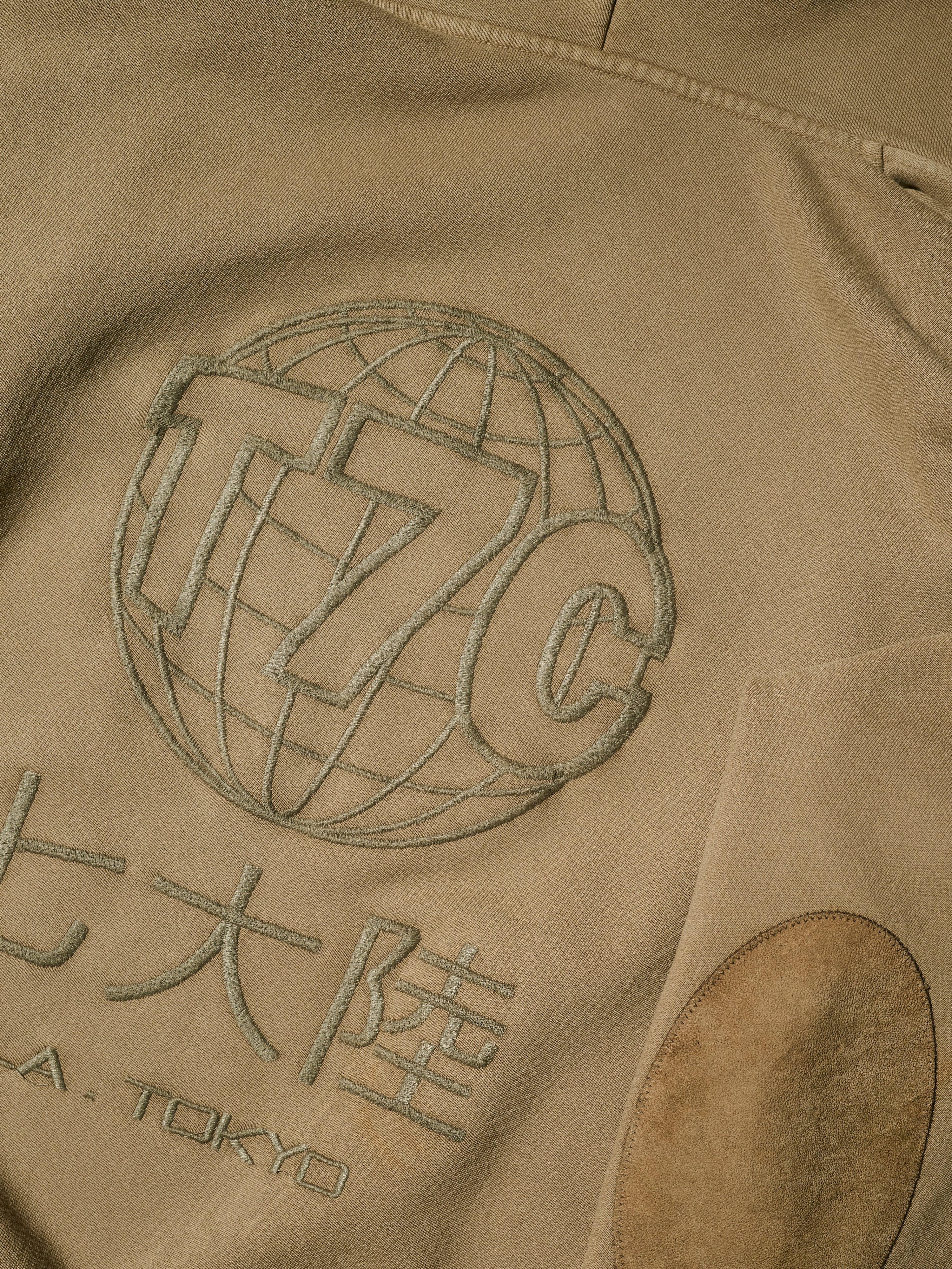 A close-up of tan fabric with embroidered details—a globe with “T7C,” Japanese characters, and “A. TOKYO” underneath. The Camden Hoodie in Desert by T7C The Seven Continents includes an oval sleeve patch and suede elbow patches for style.
