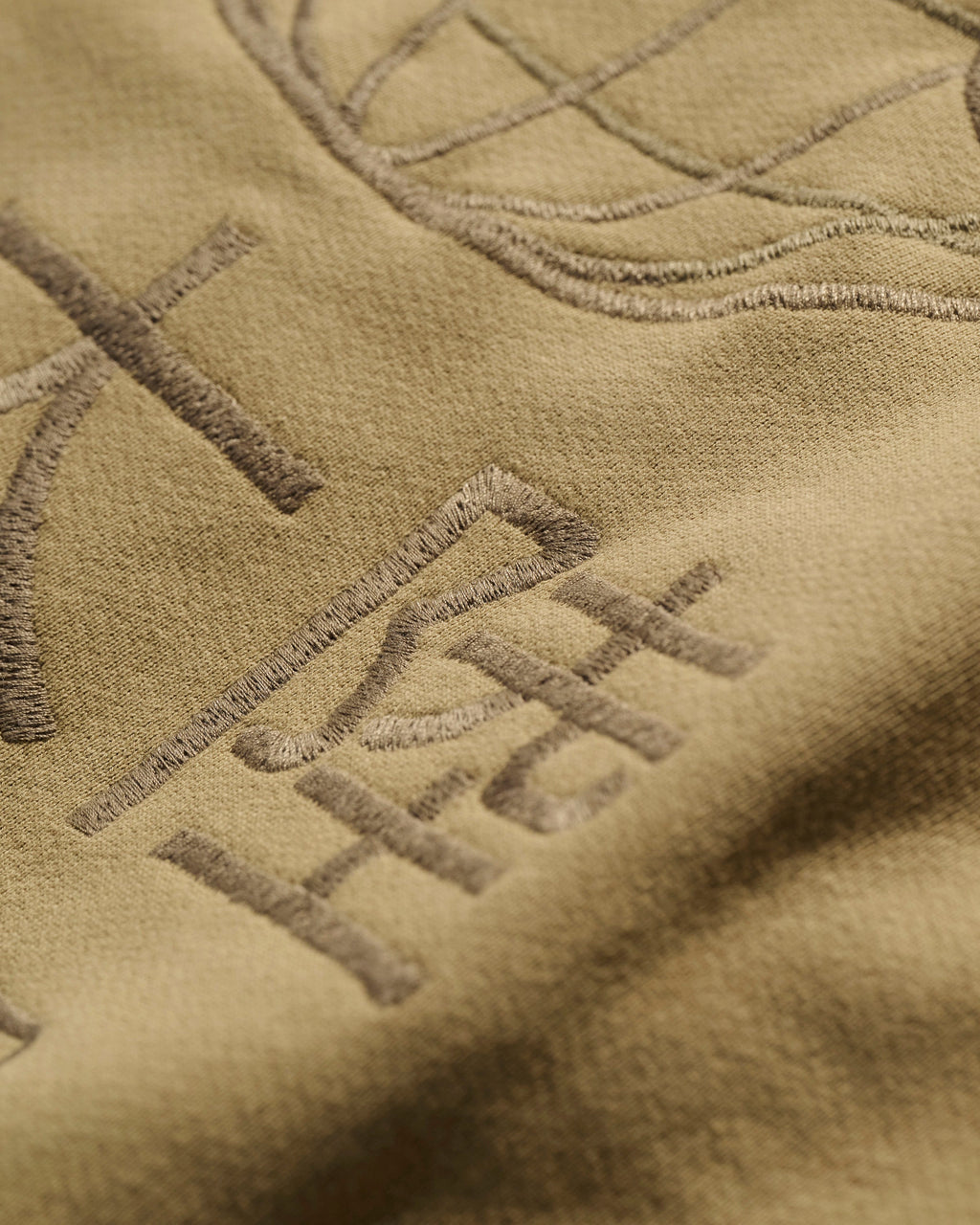 Close-up of The Camden Hoodie in Desert by T7C The Seven Continents, featuring tan fabric with raised, embroidered symbols, detailed stitching, and genuine leather accents.