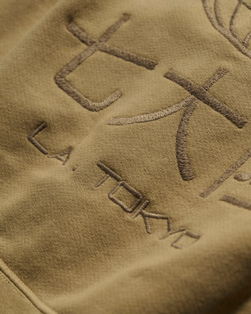 Close-up of T7C The Seven Continents The Camden Hoodie in Desert: khaki fabric with embroidered Japanese characters, L.A. TOKYO text in matching thread, and genuine leather detailing for a refined finish.