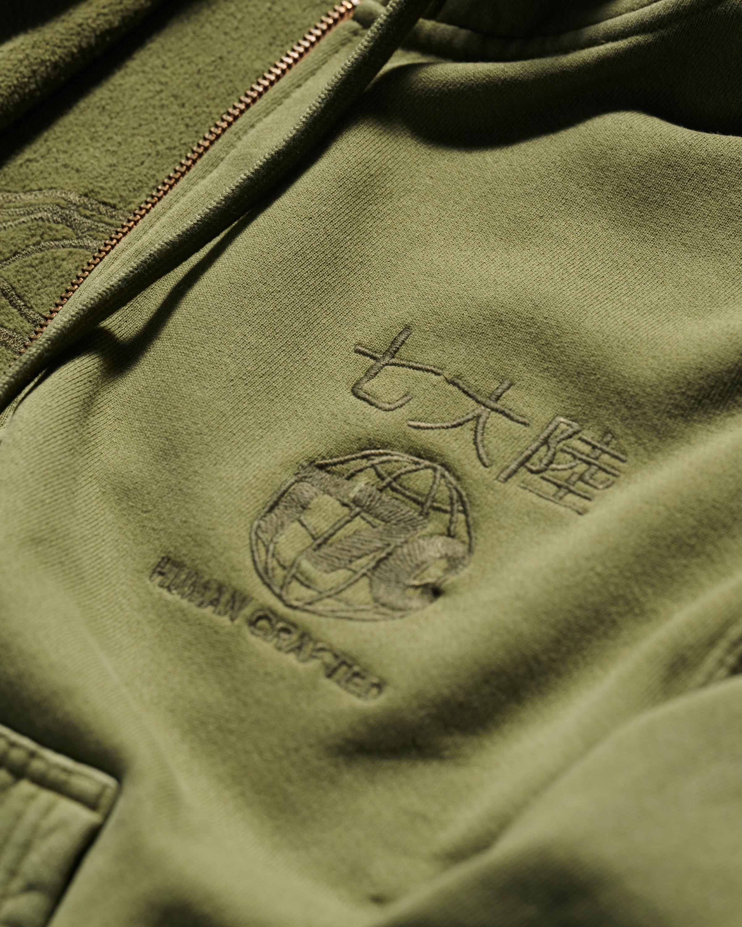 Close-up of The Camden Hoodie in Olive by T7C The Seven Continents, featuring embroidered HUMAN GRAFFITI text, a globe design, Asian characters, relaxed fit, front zipper, detailed stitching, and suede elbow patches.