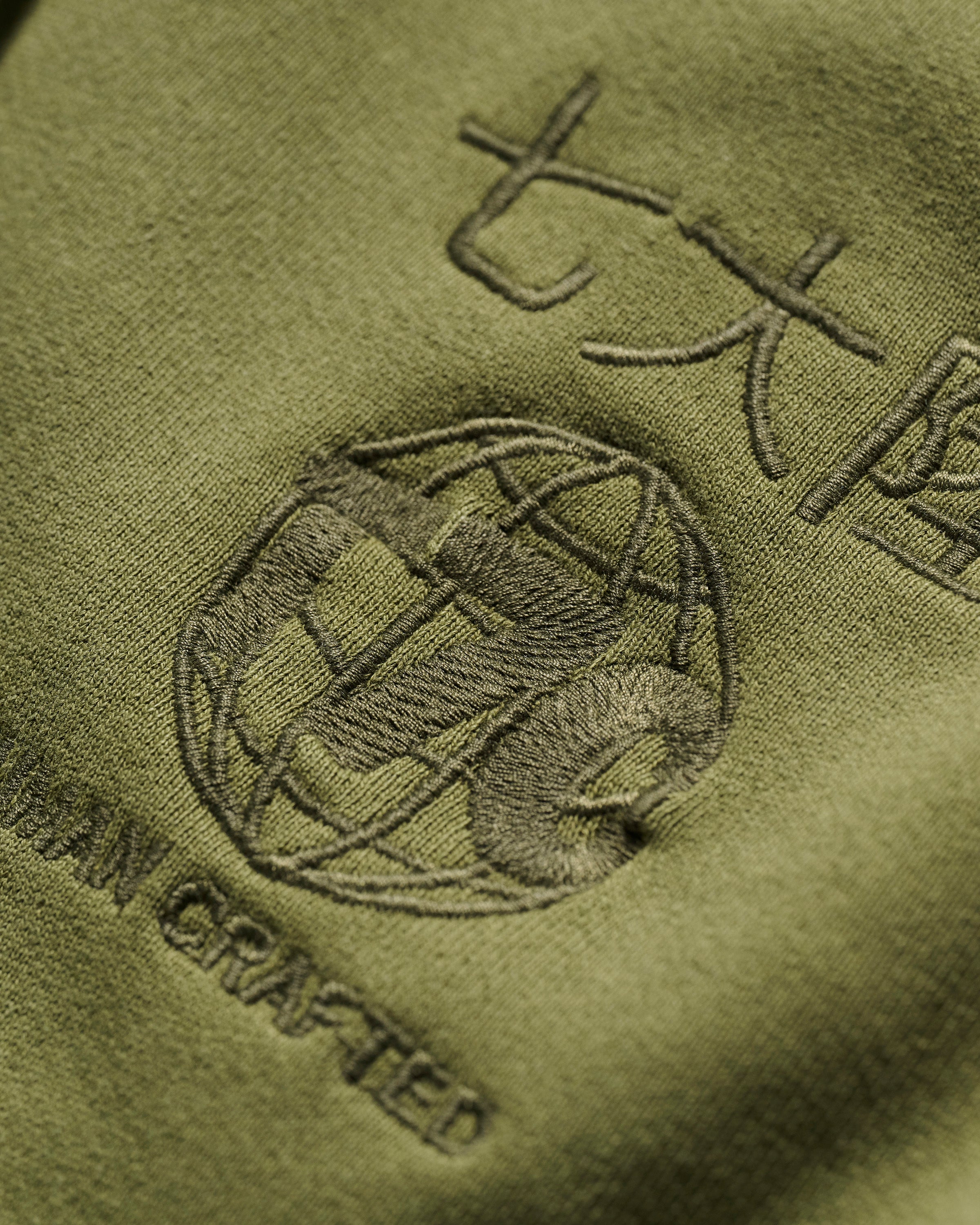 Close-up of olive green fabric from The Camden Hoodie in Olive by T7C The Seven Continents, featuring embroidered Asian characters, a globe motif, AN CRAFTED text, suede elbow patches, and detailed stitching on the textured material.