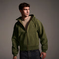 A young man in The Camden Hoodie in Olive by T7C The Seven Continents, styled over a beige tee and dark jeans, stands against a neutral background, gazing to the side with a serious look.