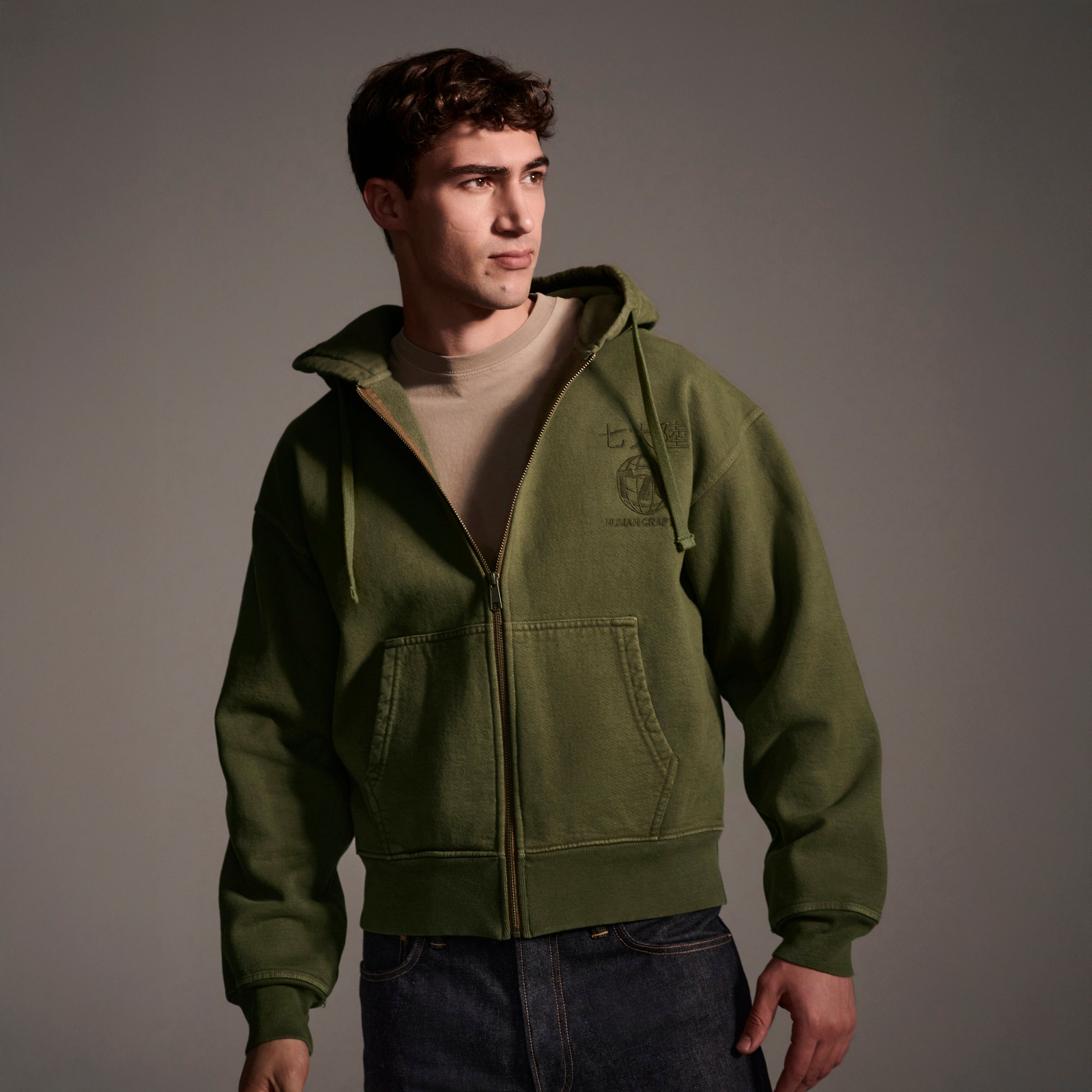 A young man in The Camden Hoodie in Olive by T7C The Seven Continents, styled over a beige tee and dark jeans, stands against a neutral background, gazing to the side with a serious look.