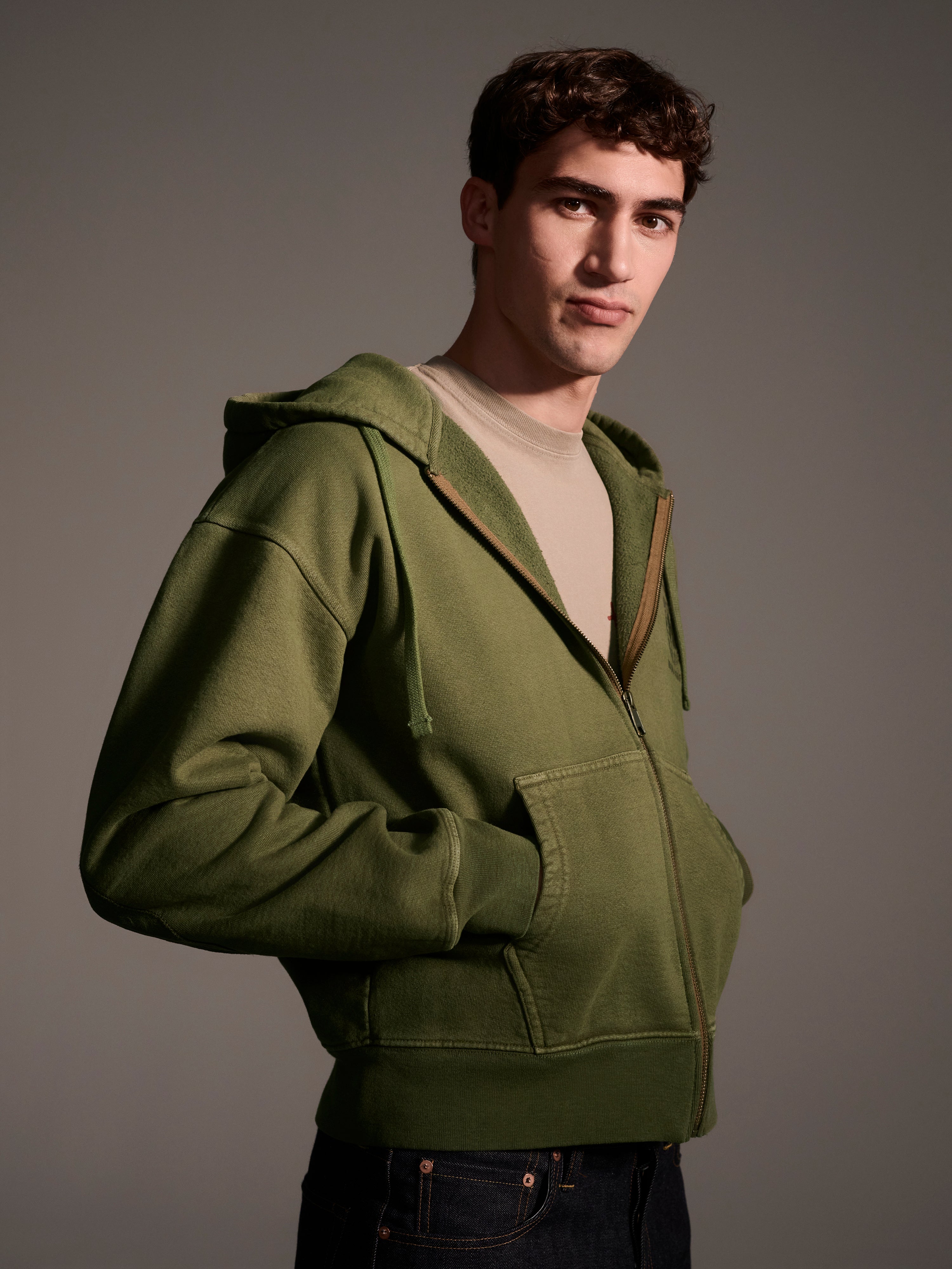 A man wears T7C The Seven Continents Camden Hoodie in Olive over a beige shirt, standing with one hand in his pocket and looking at the camera against a plain, neutral background.