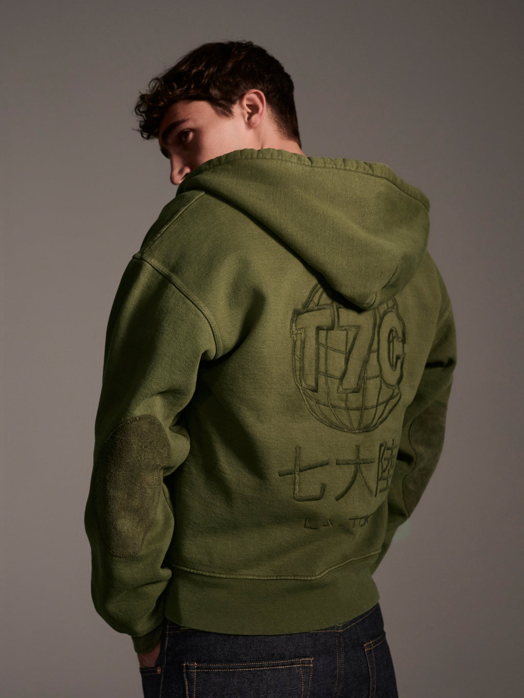 Someone stands with their back to the camera, wearing The Camden Hoodie in Olive by T7C The Seven Continents, featuring large graphic text and symbols on the back, set against a plain, neutral background.