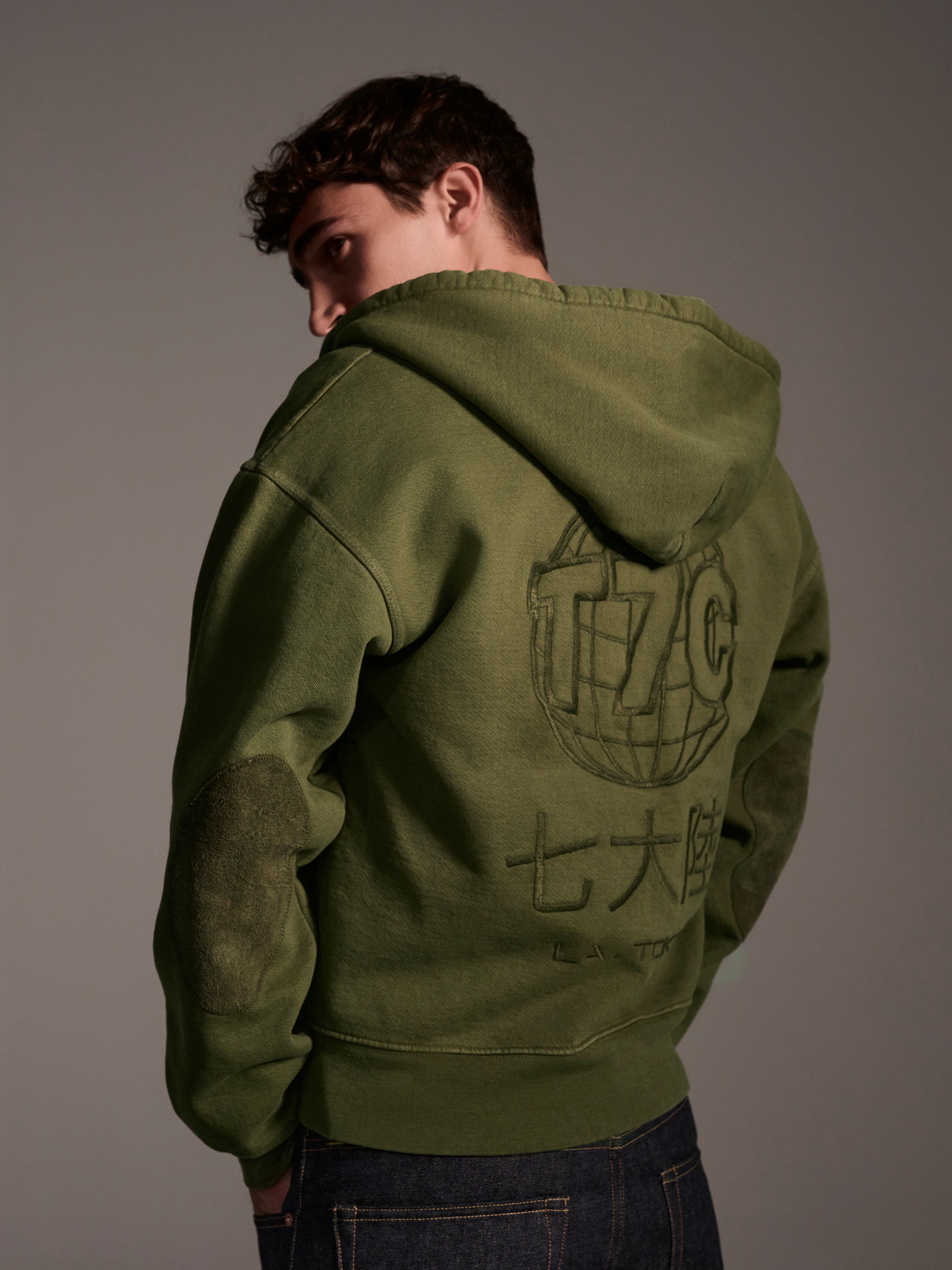 Someone stands with their back to the camera, wearing The Camden Hoodie in Olive by T7C The Seven Continents, featuring large graphic text and symbols on the back, set against a plain, neutral background.