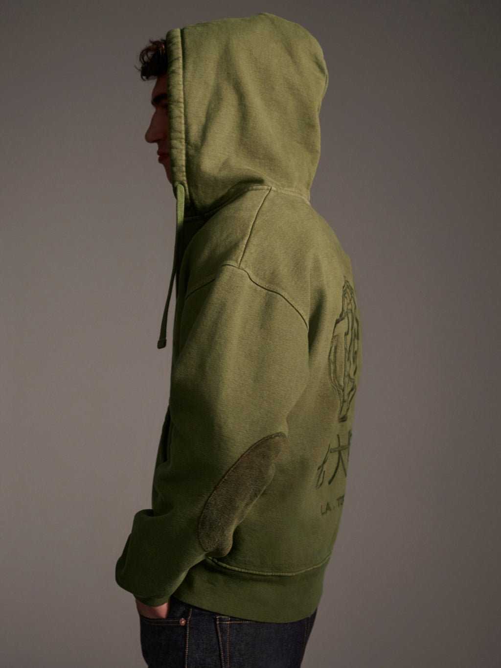 A person in The Camden Hoodie in Olive by T7C The Seven Continents stands sideways with the hood up, against a neutral background. This relaxed fit sweatshirt has a back graphic and stylish suede elbow patches.