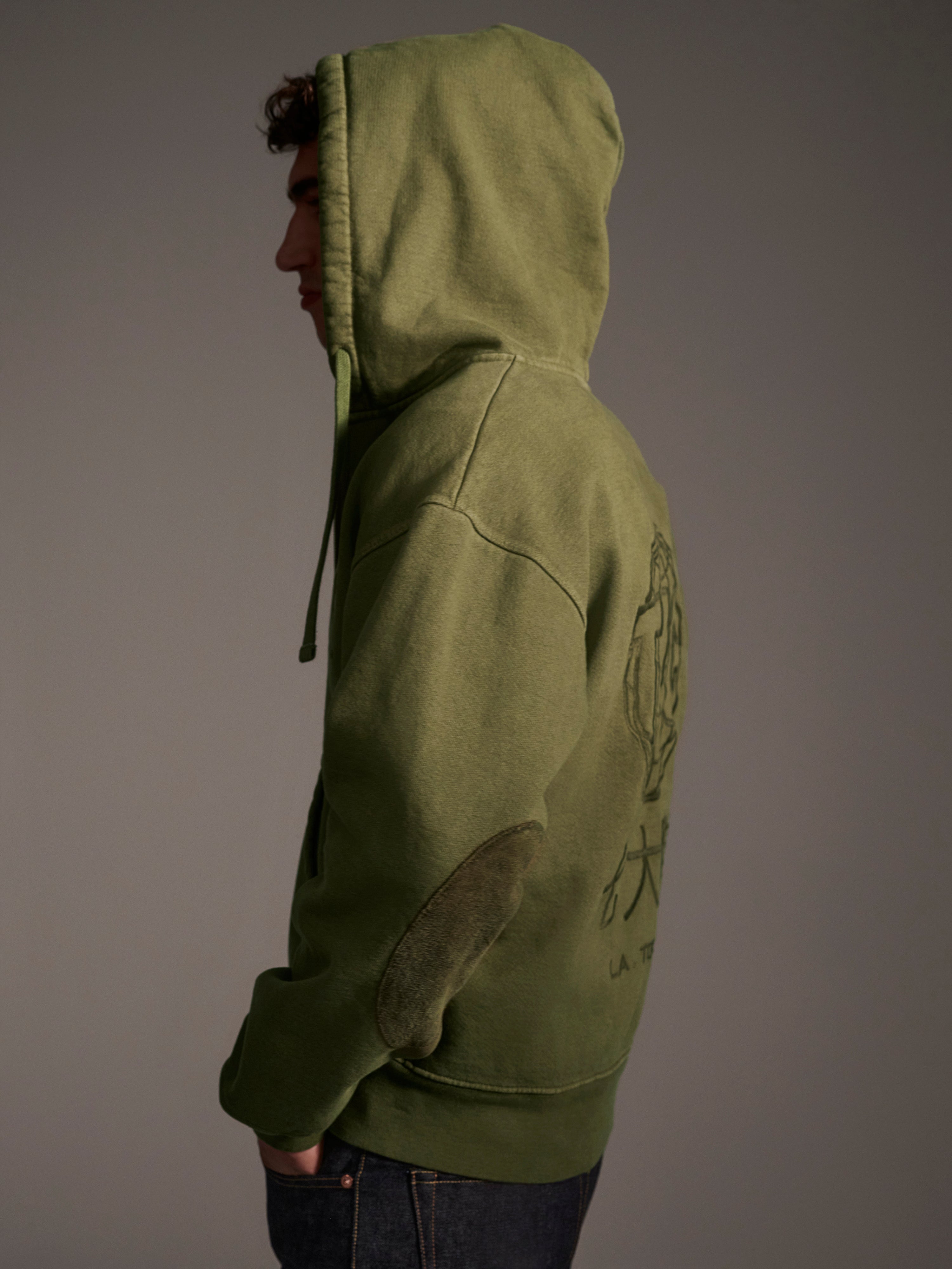 A person in The Camden Hoodie in Olive by T7C The Seven Continents stands sideways with the hood up, against a neutral background. This relaxed fit sweatshirt has a back graphic and stylish suede elbow patches.