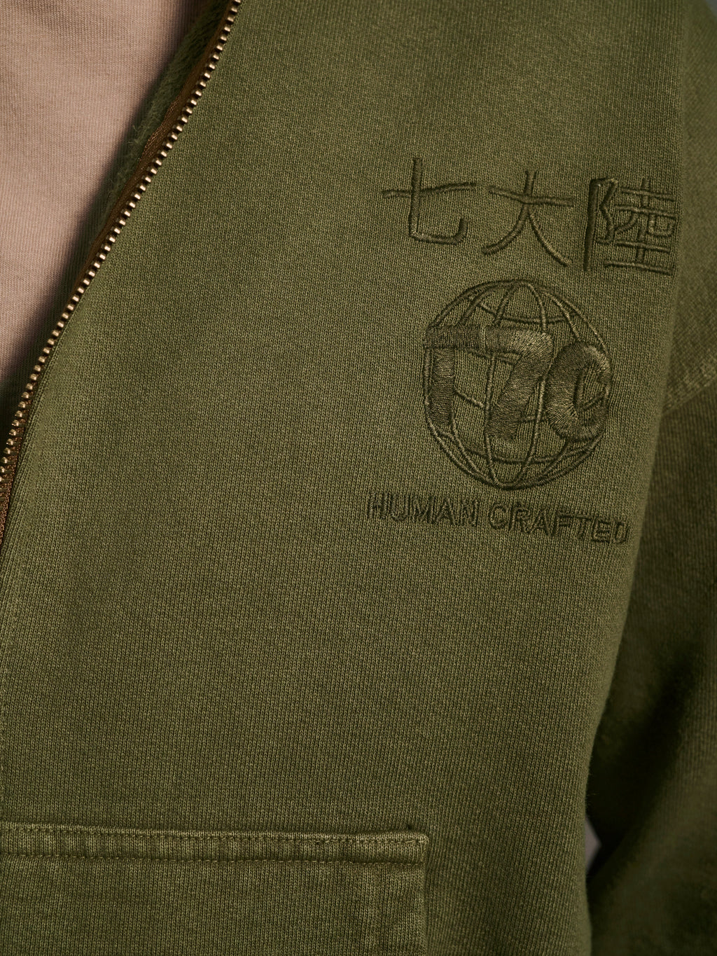 Close-up of The Camden Hoodie in Olive by T7C The Seven Continents, featuring a relaxed fit, embroidered Japanese characters, globe design, HUMAN CRAFTED text, suede elbow patches, and visible zipper and pocket.