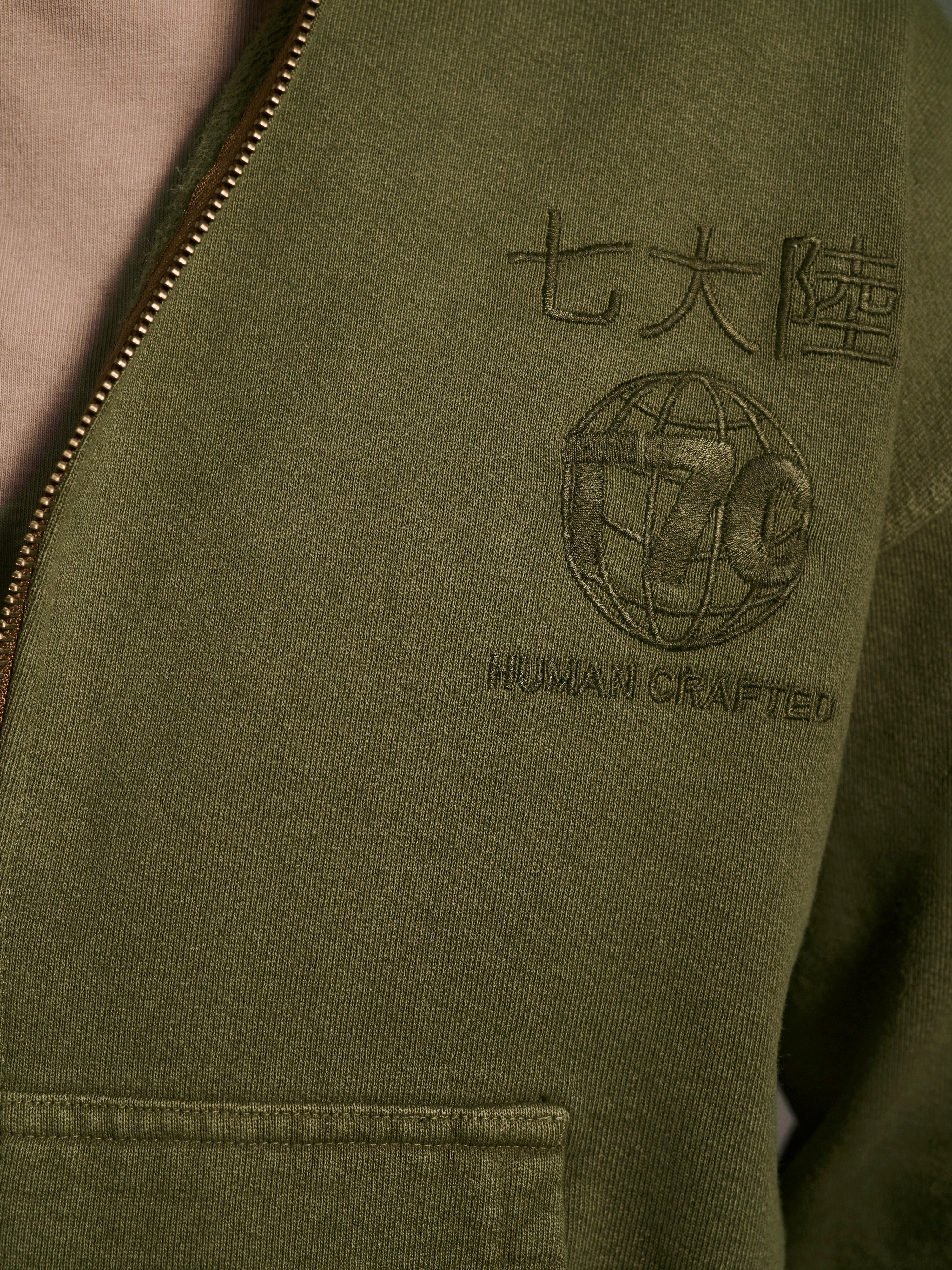 Close-up of The Camden Hoodie in Olive by T7C The Seven Continents, featuring a relaxed fit, embroidered Japanese characters, globe design, HUMAN CRAFTED text, suede elbow patches, and visible zipper and pocket.