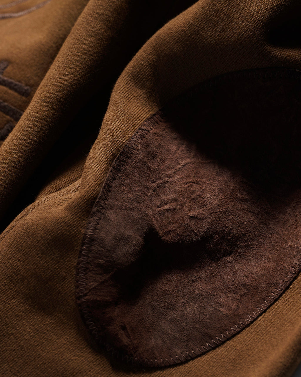 Close-up of the T7C The Seven Continents Camden Hoodie in Pecan shows its brown fabric with a darker suede elbow patch, highlighting the contrast between the smooth material and soft, textured suede.