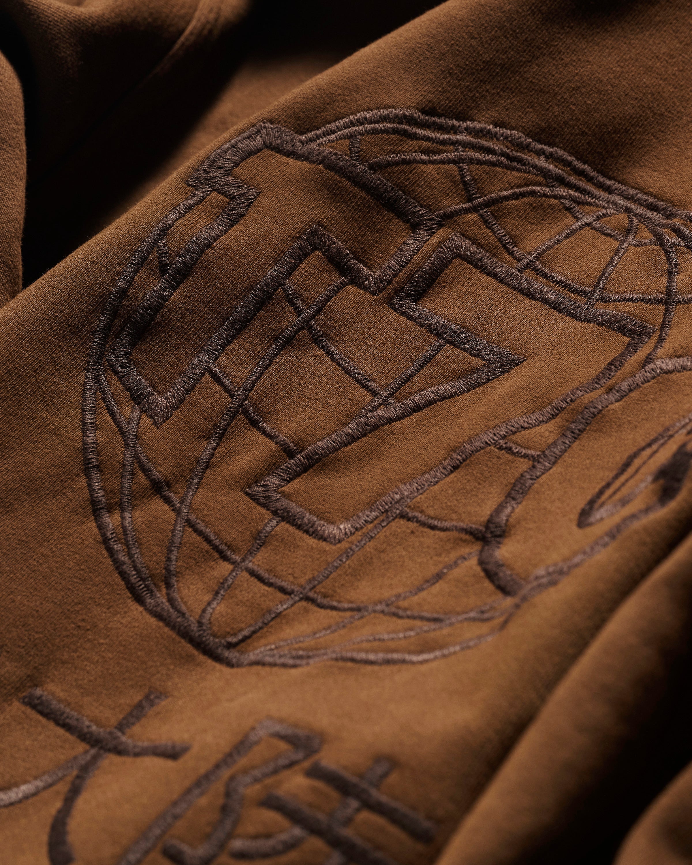 Close-up of The Camden Hoodie in Pecan by T7C The Seven Continents, showcasing brown fabric with an embroidered globe, geometric designs, stylized characters, and suede elbow patches for a relaxed, detailed look.