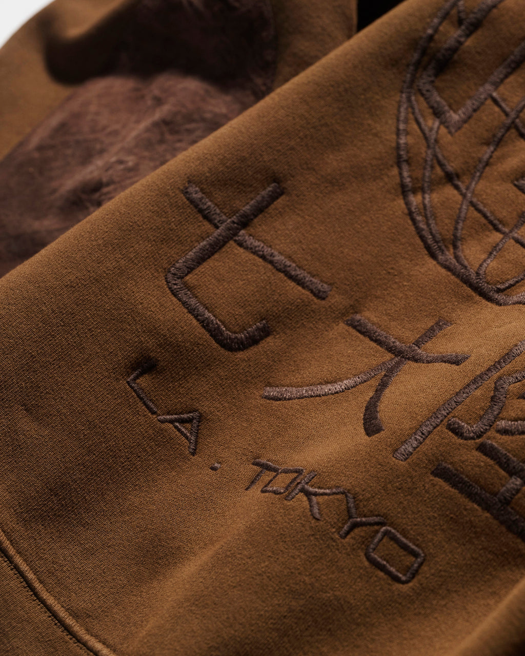 A close-up of The Camden Hoodie in Pecan by T7C The Seven Continents shows brown fabric with L.A. TOKYO and large Japanese characters embroidered beside a partial stitched globe, all in a darker brown thread for subtle detail.