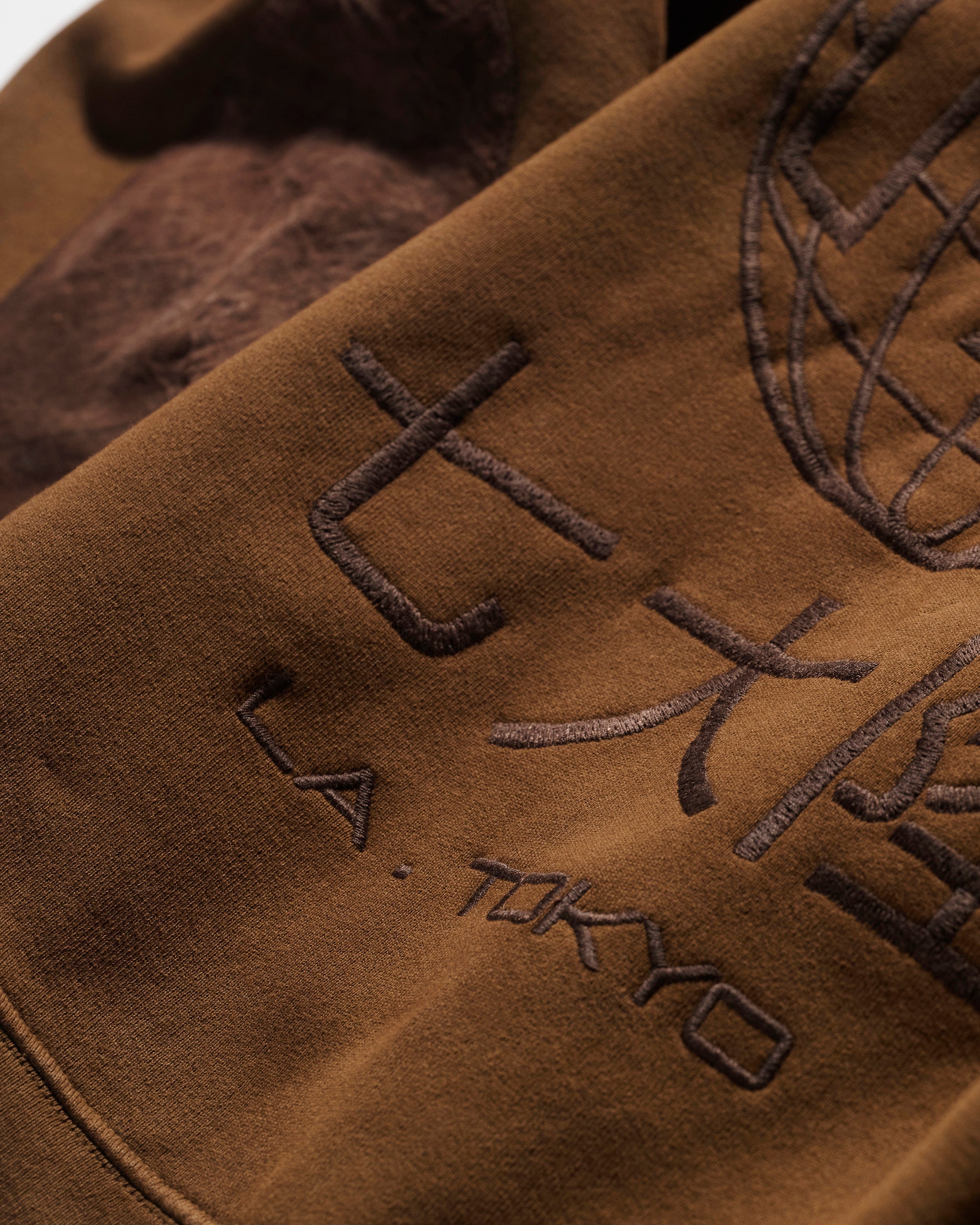 A close-up of The Camden Hoodie in Pecan by T7C The Seven Continents shows brown fabric with L.A. TOKYO and large Japanese characters embroidered beside a partial stitched globe, all in a darker brown thread for subtle detail.
