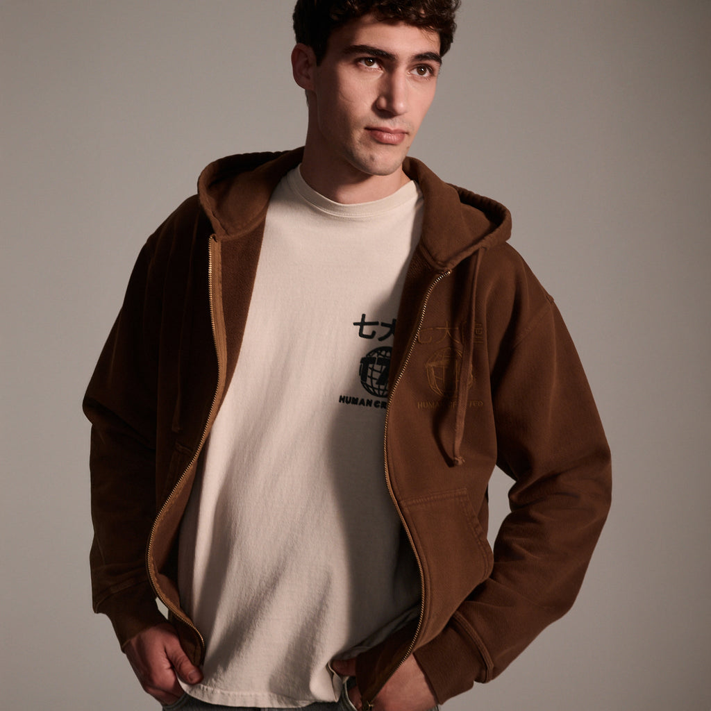 A young man stands with hands in pockets, wearing T7C The Seven Continents Camden Hoodie in Pecan—a relaxed fit zip-up with suede elbow patches—over a light T-shirt with Japanese characters, set against a plain background.