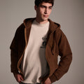 A young man stands with hands in pockets, wearing T7C The Seven Continents Camden Hoodie in Pecan—a relaxed fit zip-up with suede elbow patches—over a light T-shirt with Japanese characters, set against a plain background.