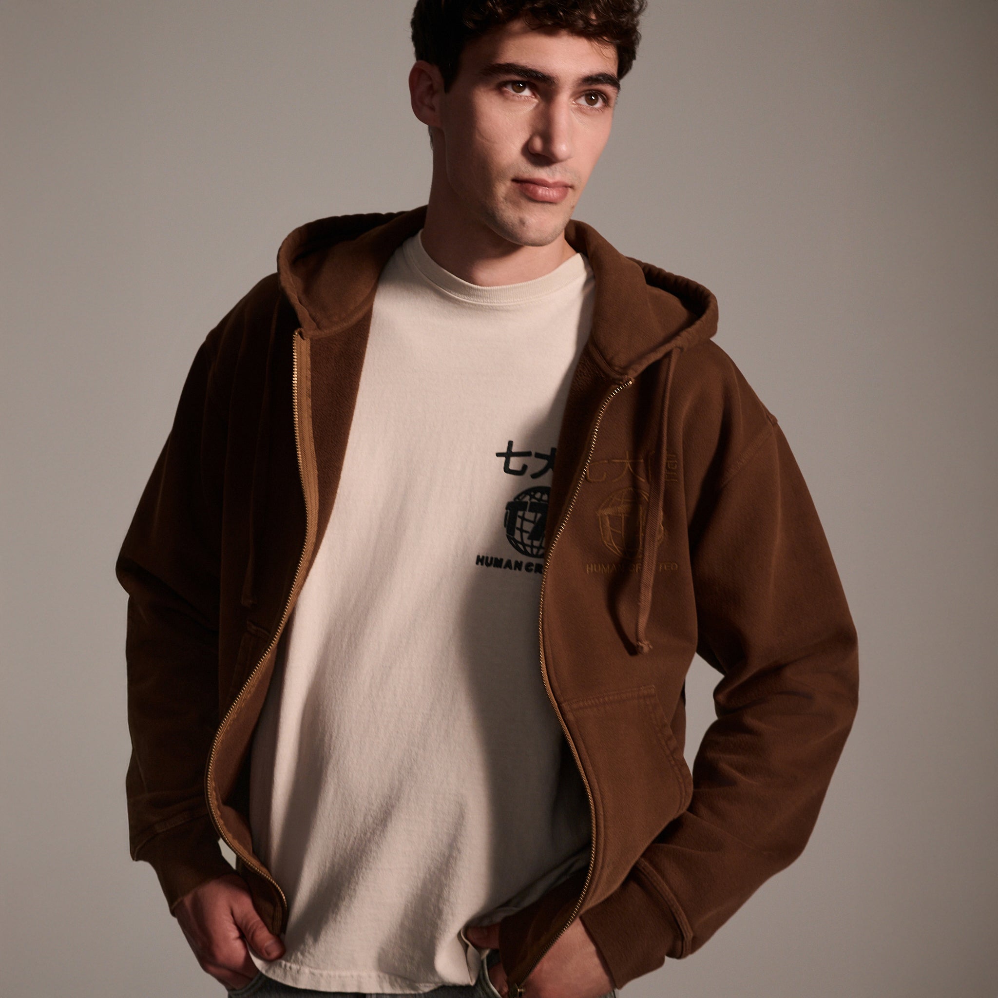 A young man stands with hands in pockets, wearing T7C The Seven Continents Camden Hoodie in Pecan—a relaxed fit zip-up with suede elbow patches—over a light T-shirt with Japanese characters, set against a plain background.