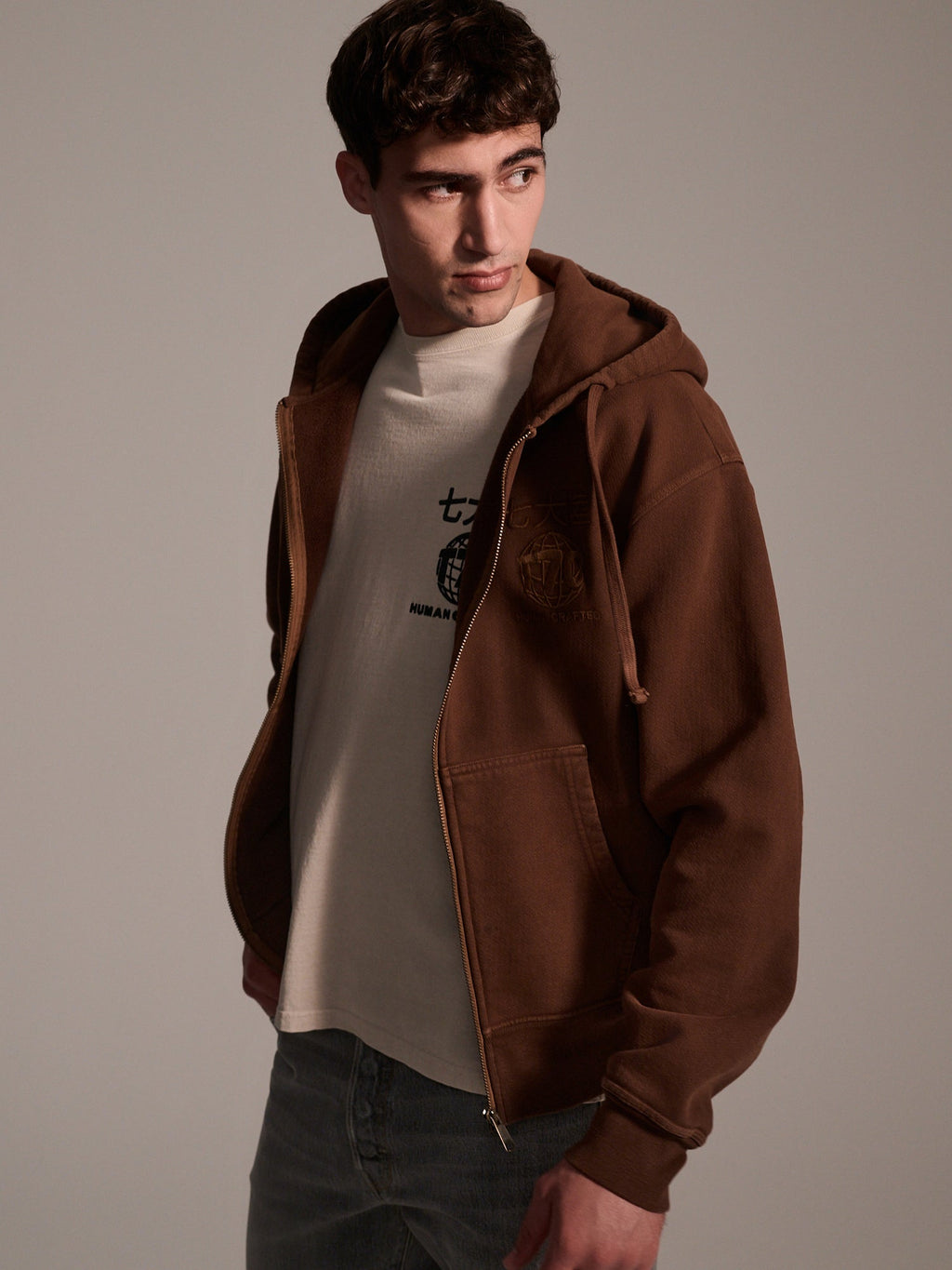 A young man with short brown hair wears The Camden Hoodie in Pecan by T7C The Seven Continents over a light graphic tee and faded jeans, glancing left against a neutral background.