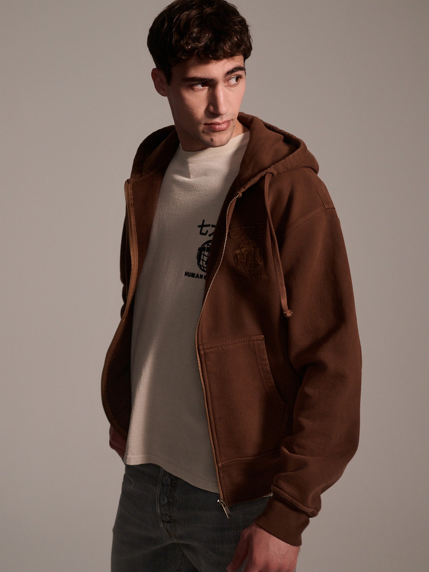 A young man with short brown hair wears The Camden Hoodie in Pecan by T7C The Seven Continents over a light graphic tee and faded jeans, glancing left against a neutral background.