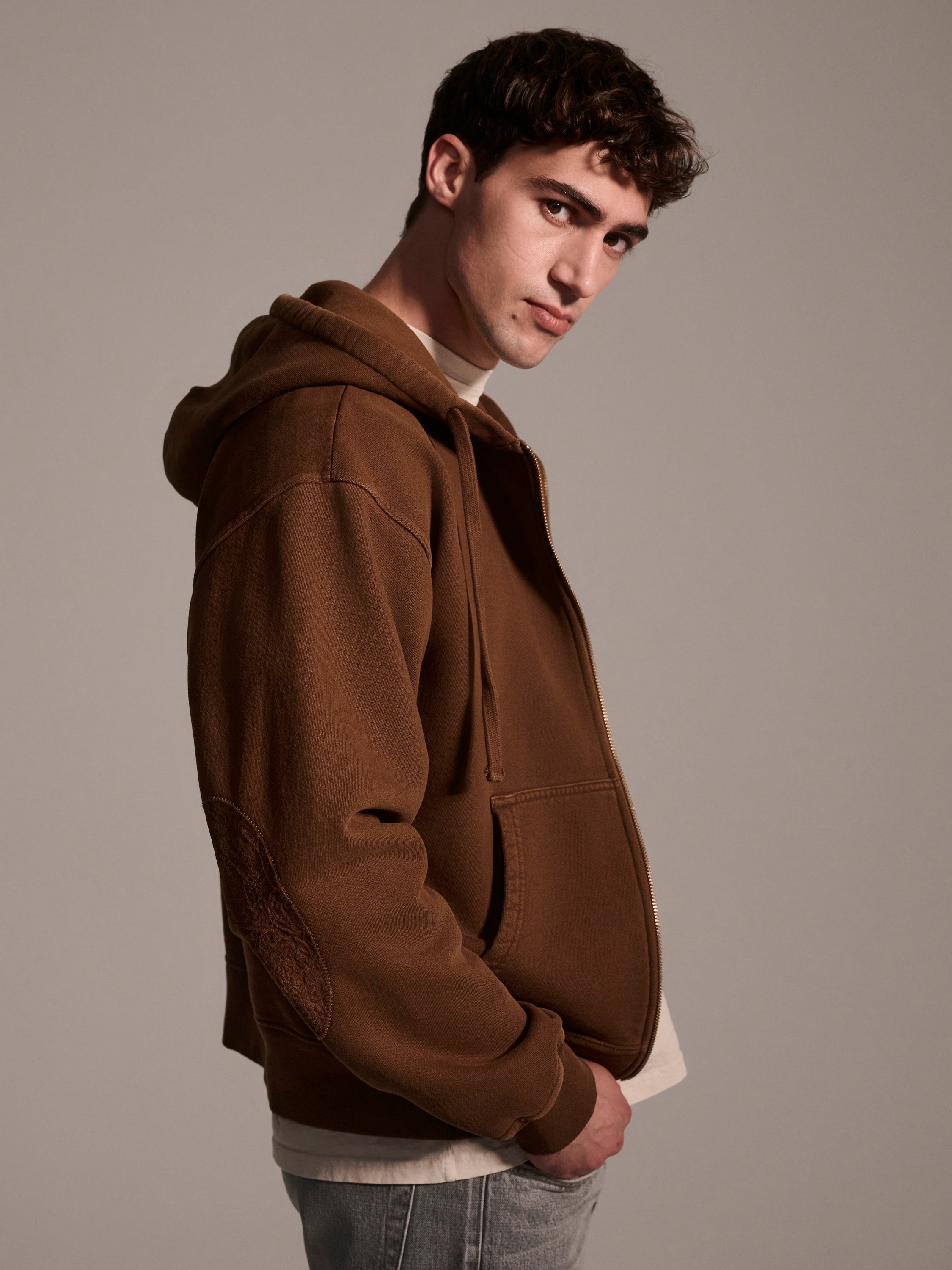 A young man models The Camden Hoodie in Pecan by T7C The Seven Continents over a white shirt, facing left with his head turned slightly to the camera, posing against a plain, neutral background.