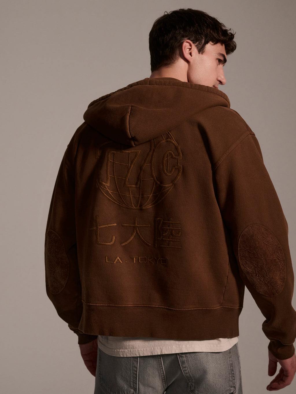 A man stands turned slightly to the side, wearing The Camden Hoodie in Pecan by T7C The Seven Continents. This relaxed fit brown hoodie features an embroidered globe, stylized letters, Japanese characters, suede elbow patches, and L.A. TOKYO on the back. He pairs it with light jeans.
