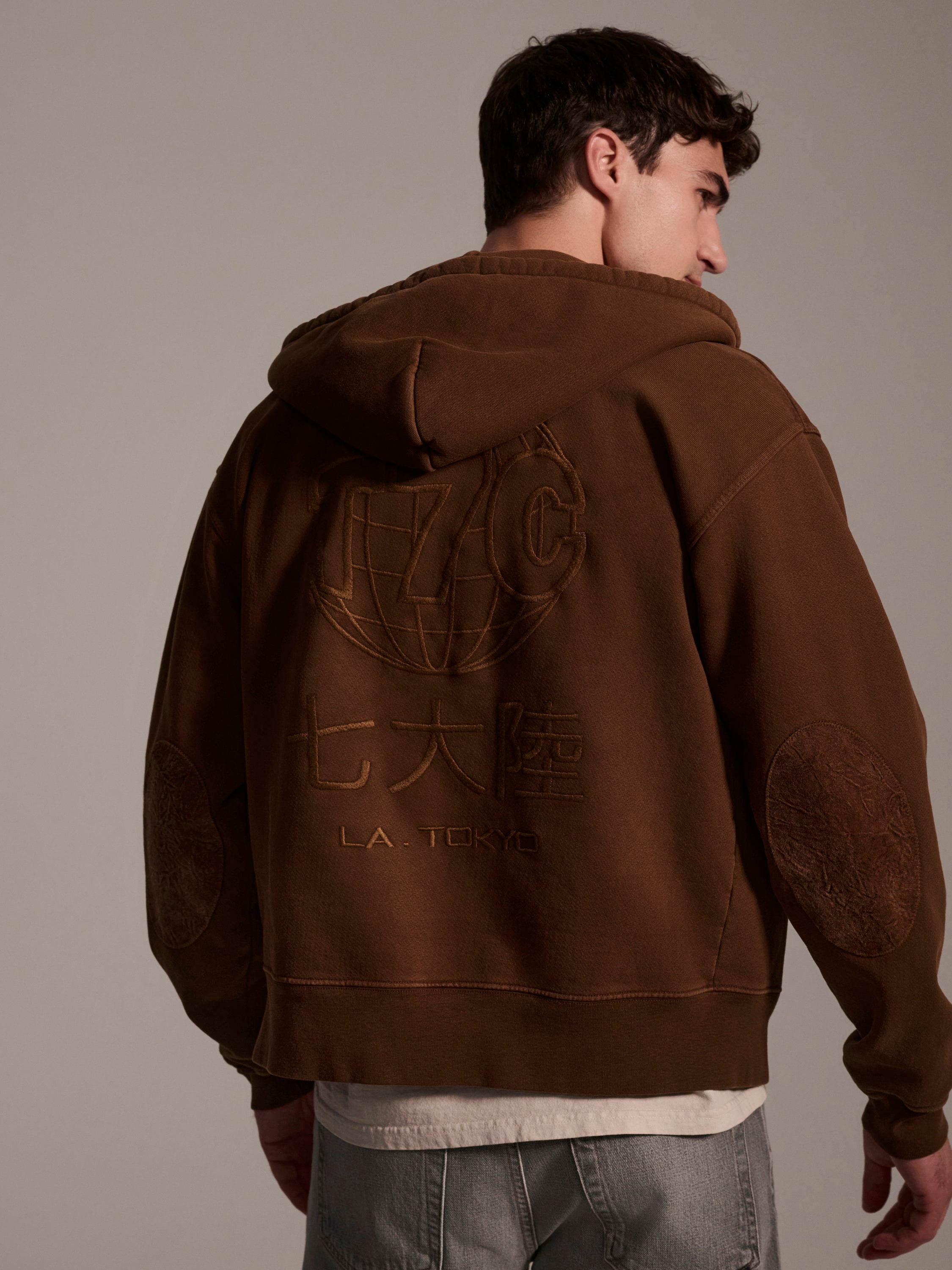 A man stands turned slightly to the side, wearing The Camden Hoodie in Pecan by T7C The Seven Continents. This relaxed fit brown hoodie features an embroidered globe, stylized letters, Japanese characters, suede elbow patches, and L.A. TOKYO on the back. He pairs it with light jeans.