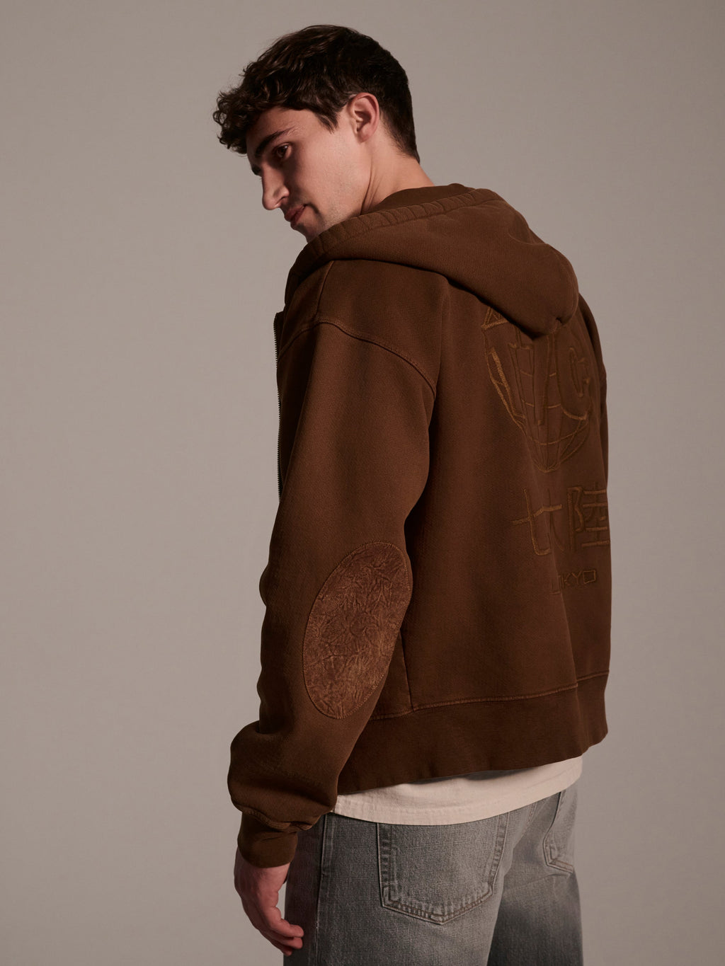 A young man with short curly hair models the T7C The Seven Continents Camden Hoodie in Pecan, featuring suede elbow patches and embossed back designs, paired with light gray jeans as he looks over his shoulder against a plain background.