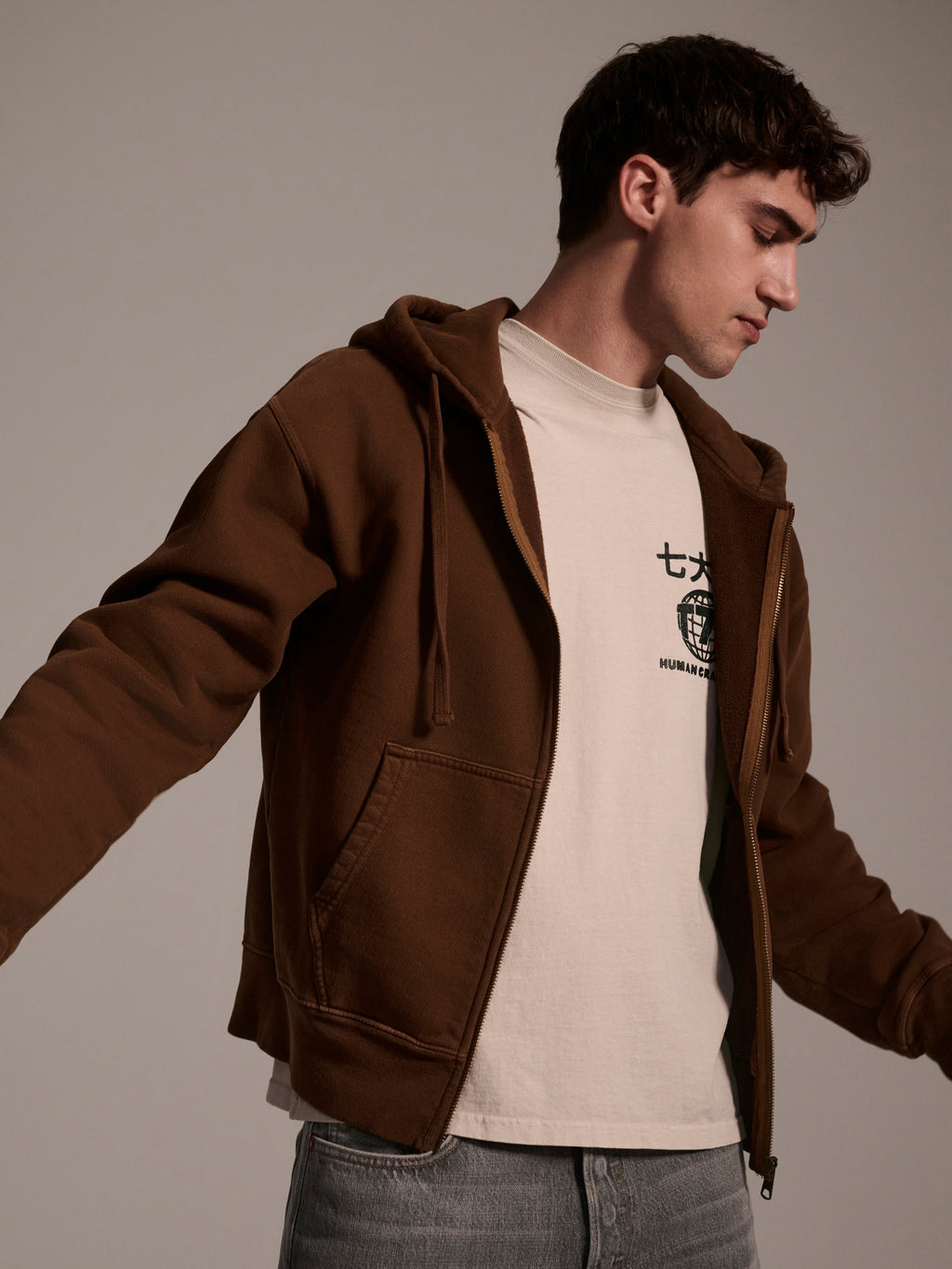 A young man wears The Camden Hoodie in Pecan by T7C The Seven Continents—relaxed fit with suede elbow patches—over a cream graphic tee and blue jeans, standing with arms slightly extended against a plain background.