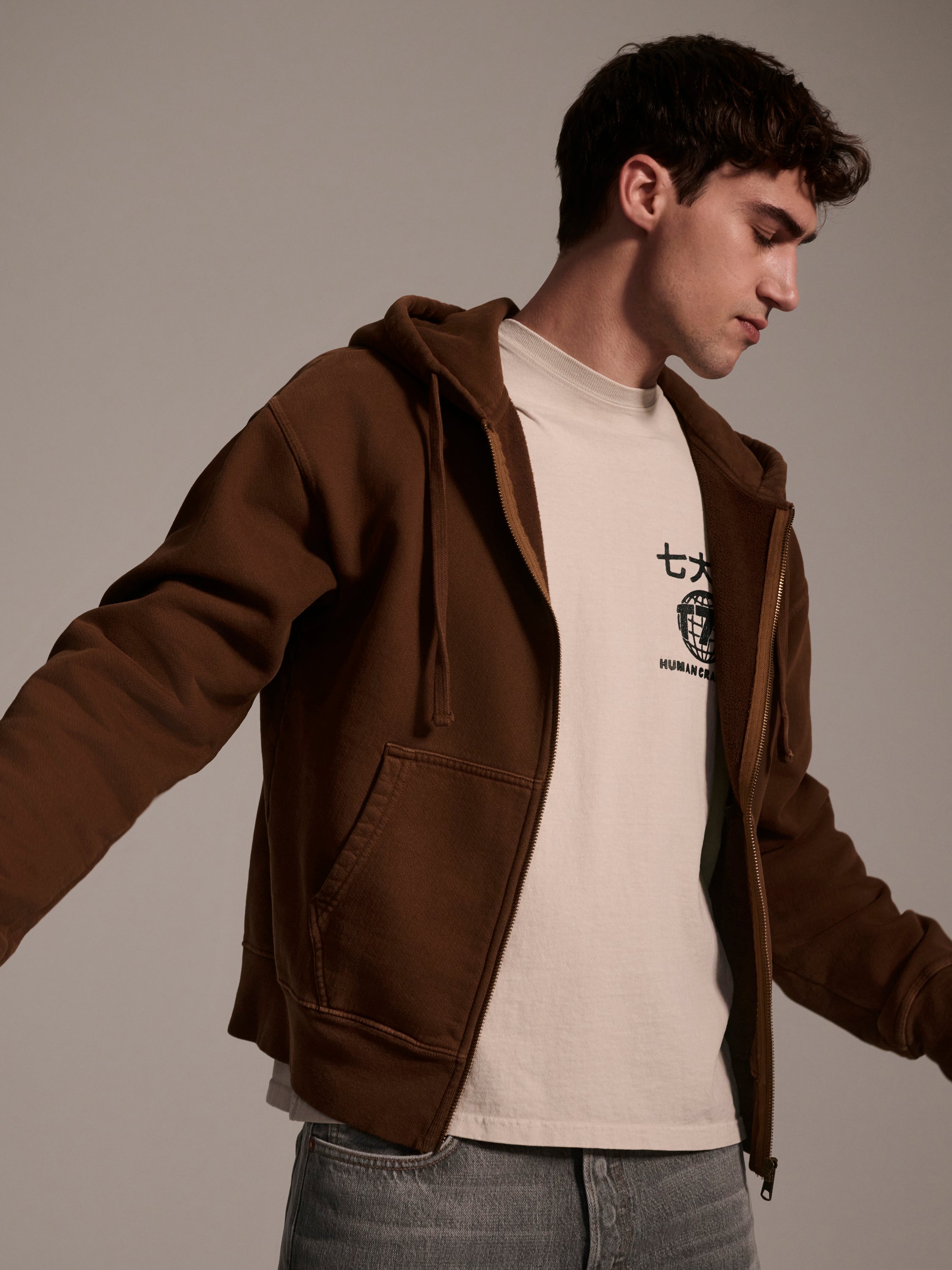 A young man wears The Camden Hoodie in Pecan by T7C The Seven Continents—relaxed fit with suede elbow patches—over a cream graphic tee and blue jeans, standing with arms slightly extended against a plain background.