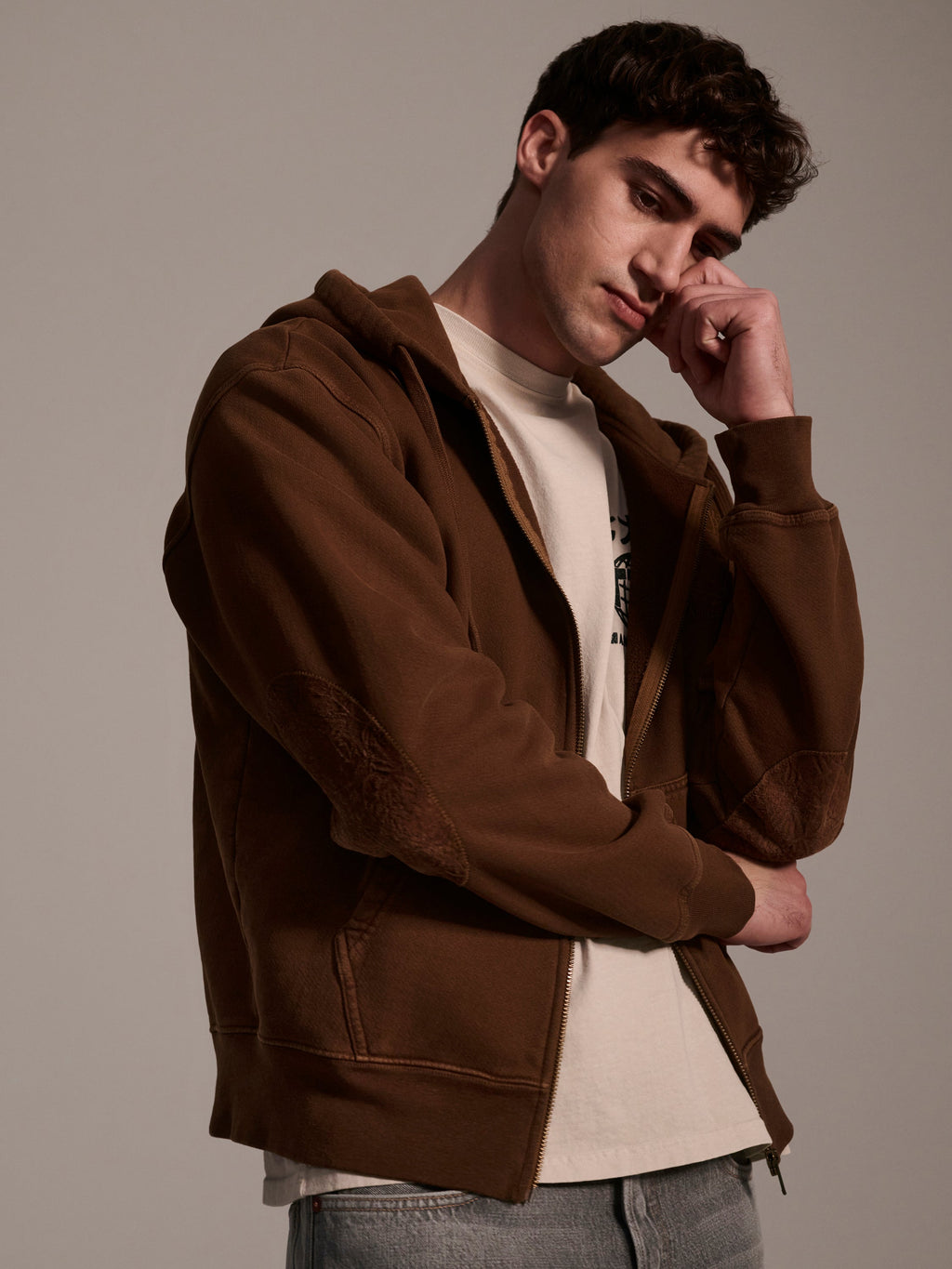 A young man with short, dark hair wears The Camden Hoodie in Pecan by T7C The Seven Continents over a white t-shirt and light jeans, standing thoughtfully with one hand touching his face and the other arm crossed.