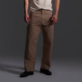 Only the lower half of a person is visible, wearing T7Cs The Fred Pant in Camel Stripe—relaxed-fit carpenter pants made from Japanese mill fabric—paired with a light-colored t-shirt and black shoes, set against a dark background.