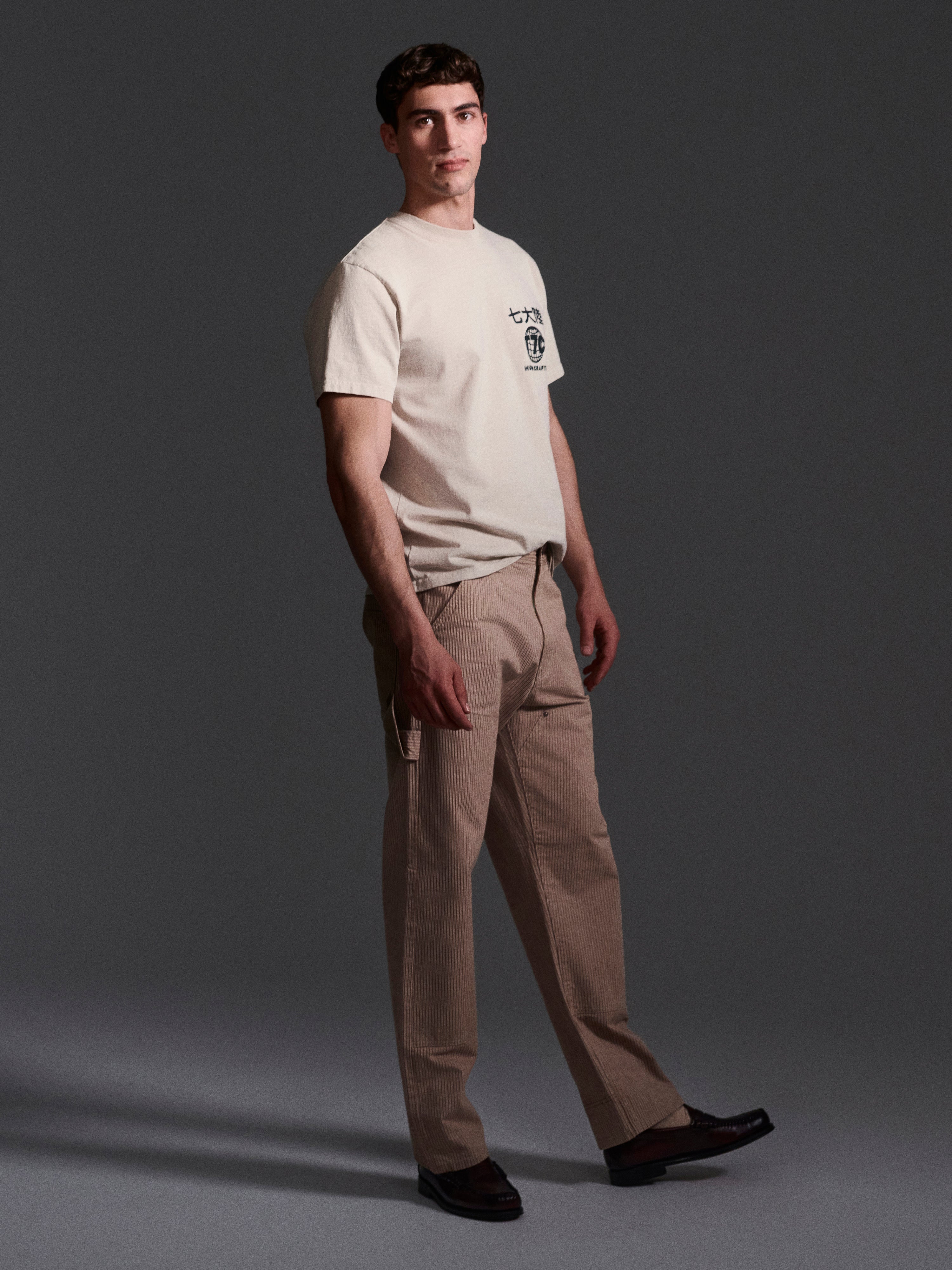 A man stands against a dark background, wearing a beige T-shirt with a small chest graphic, T7Cs The Fred Pant in Camel Stripe made from Japanese mill fabric in a relaxed fit, and black shoes. He faces slightly to the side with a neutral expression.