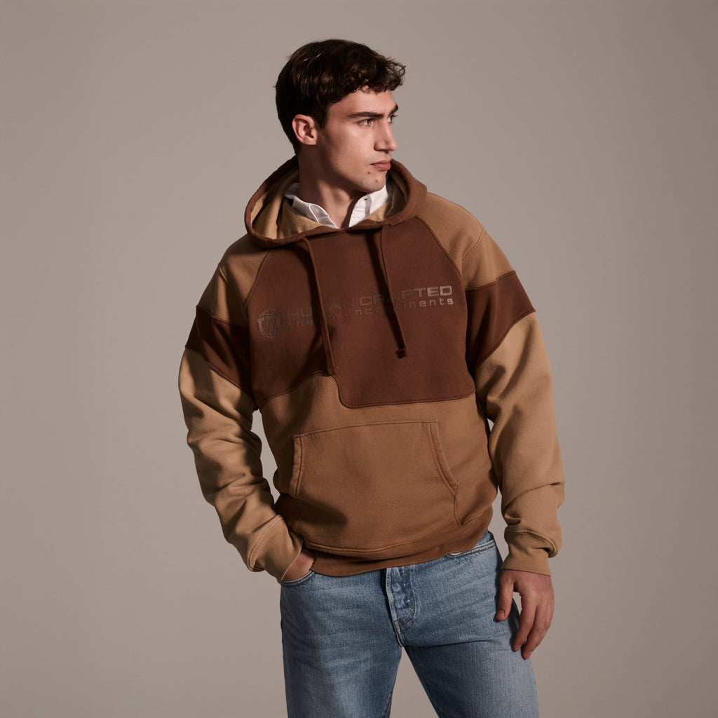 A man wears the T7C The Seven Continents Hunter Hoodie in Terra over a white collared shirt and blue jeans, standing against a plain background with one hand in his pocket, looking to his left.