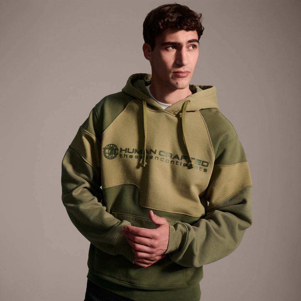 A young man stands against a neutral background, wearing T7C The Seven Continents Hunter Hoodie in Verde, featuring “Human Crafted the 7 Continents” on the front and camo-inspired seams, hands lightly clasped as he looks to the side.