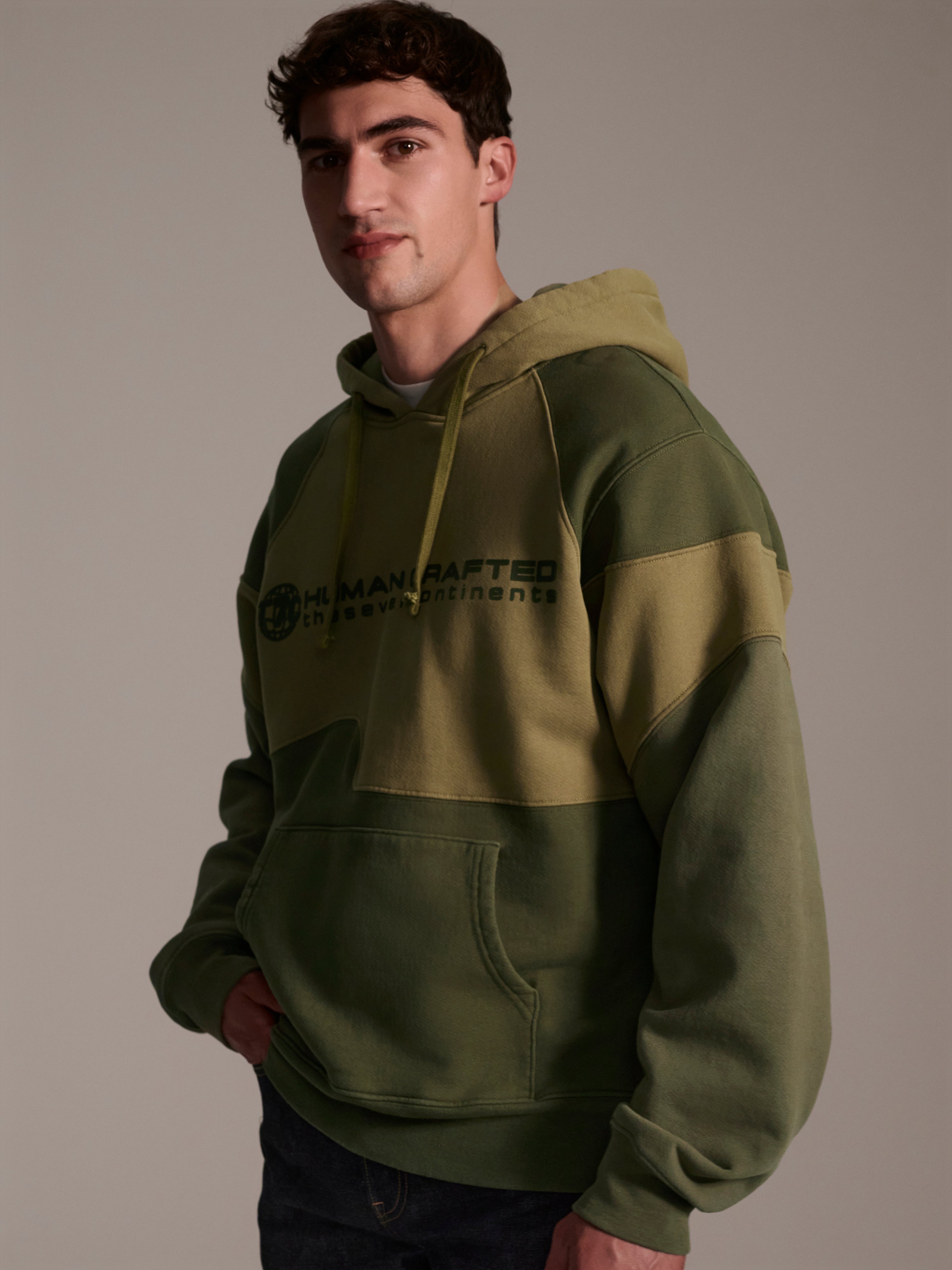 A profile image of a male model wearing a color blocked pull-over hoodie in green tones featuring a printed logo 