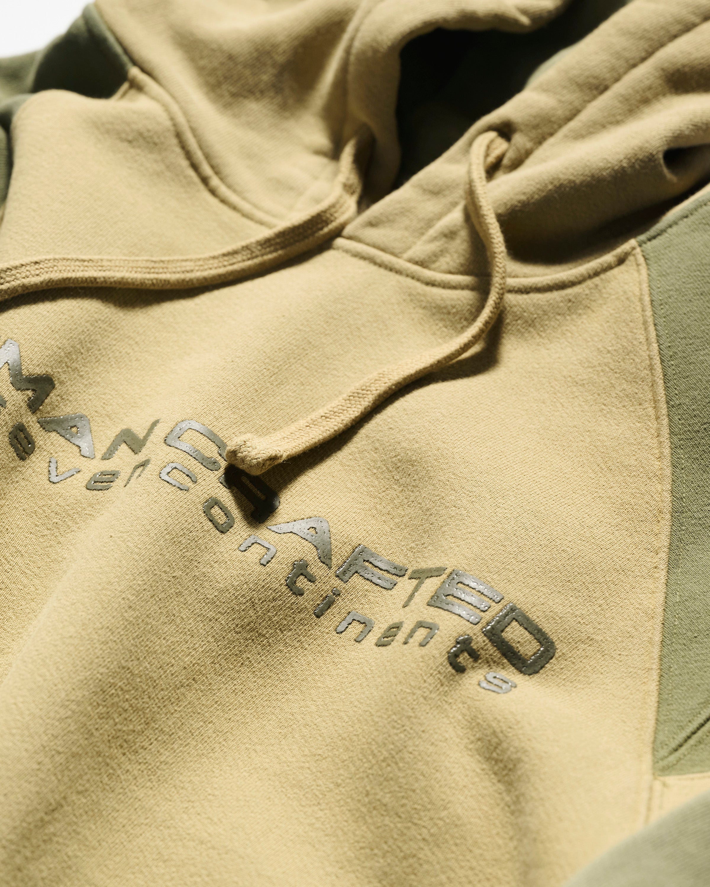 A close-up of The Hunter Hoodie in Verde by T7C The Seven Continents, featuring a two-tone beige and olive green design with MANCRAFTED overcontinents in raised metallic letters, plus camo-inspired seam and drawstring details.
