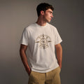 A young man wears the T7C PRE-ORDER: THE KNOXS TEE WITH MERIDIAN PRINT, featuring an eye graphic and text, with his hands in tan pants pockets, looking to the side against a neutral background.
