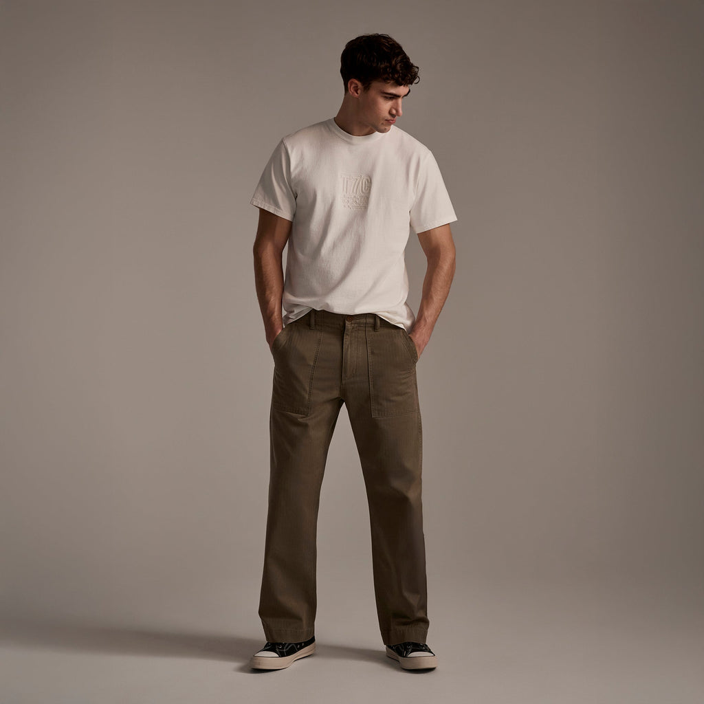 A man stands against a plain background, wearing a white t-shirt and T7C’s The Liam Military Pant in Olive Drab with black sneakers. His hands are in his pockets and he looks down to his left.